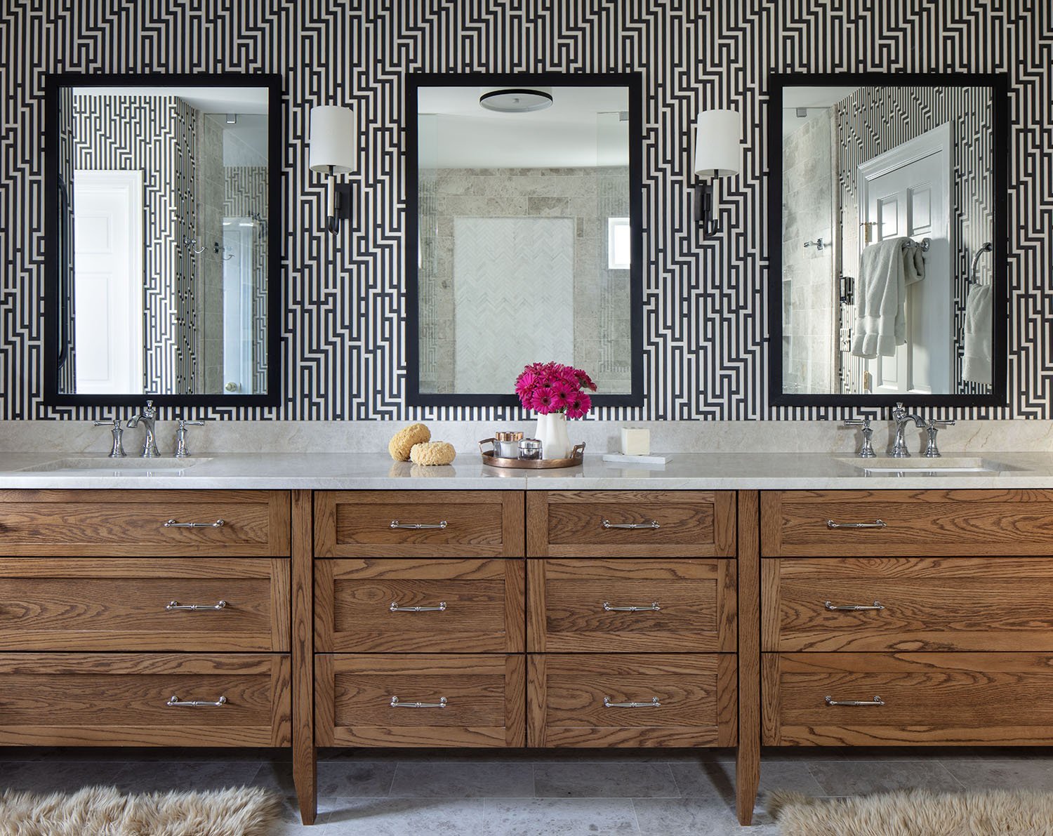 A modern bathroom with a wooden vanity, three mirrors, and black and white geometric wallpaper. The vanity has a white countertop with a pink flower vase, soap, and bath poufs.