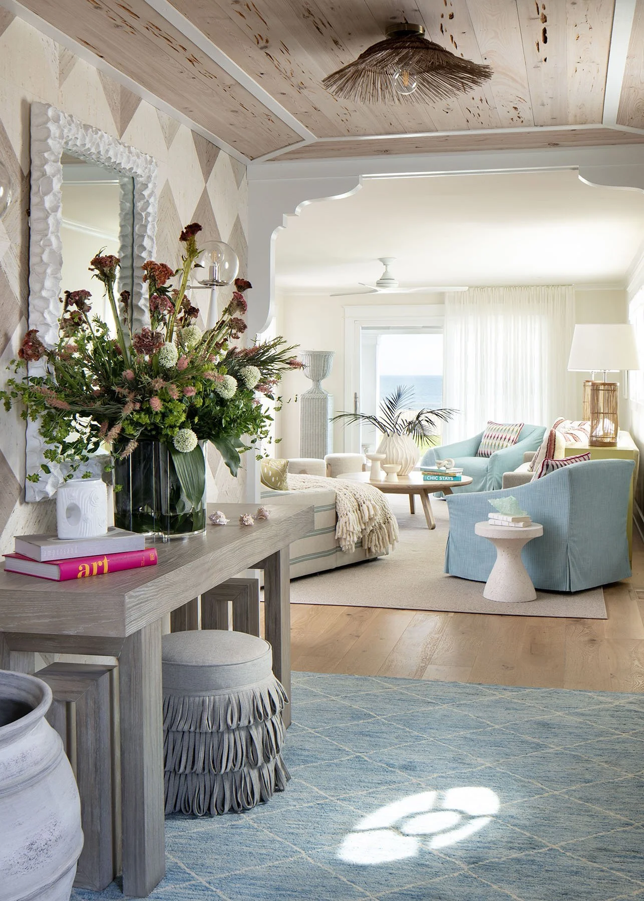 Living room with light blue armchair, a wooden table, white curtains, and a large window overlooking the ocean, decorated in a coastal style.
