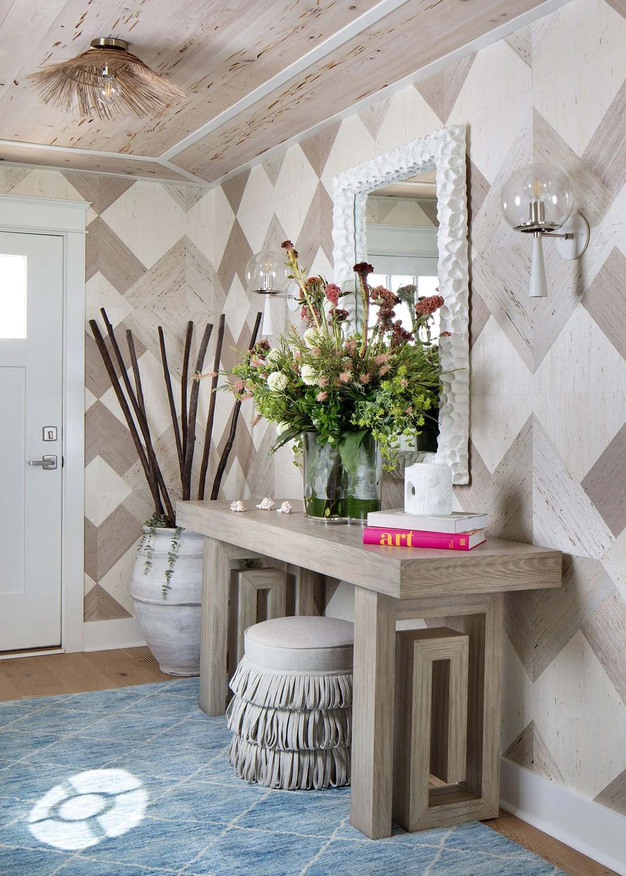 A decorated foyer with a patterned beige and white wall, a wooden console table holding a large floral arrangement, books, and a white decorative object. There is a white upholstered stool with a striped fabric cover, a large white ceramic pot with t