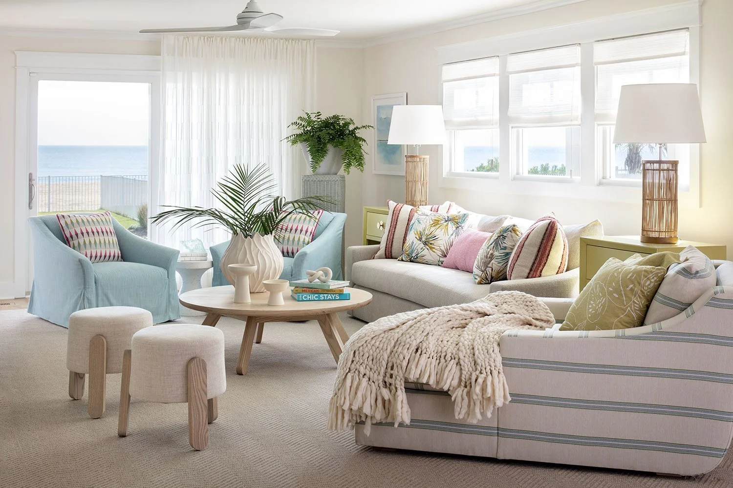 Coastal living room with large windows, white walls, pastel furniture, a sofa decorated with colorful pillows, two blue armchairs with patterned pillows, a coffee table with a large vase and books, and a view of the ocean outside.