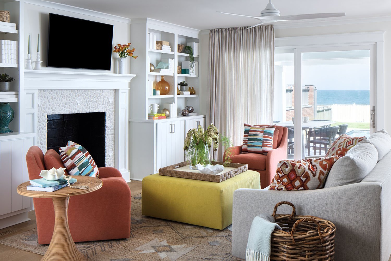 A bright living room with a view of the ocean through glass sliding doors. The room features a white fireplace with a flat-screen TV above, colorful armchairs, a white sofa, a yellow ottoman, and various decorative items on built-in shelves and a sid