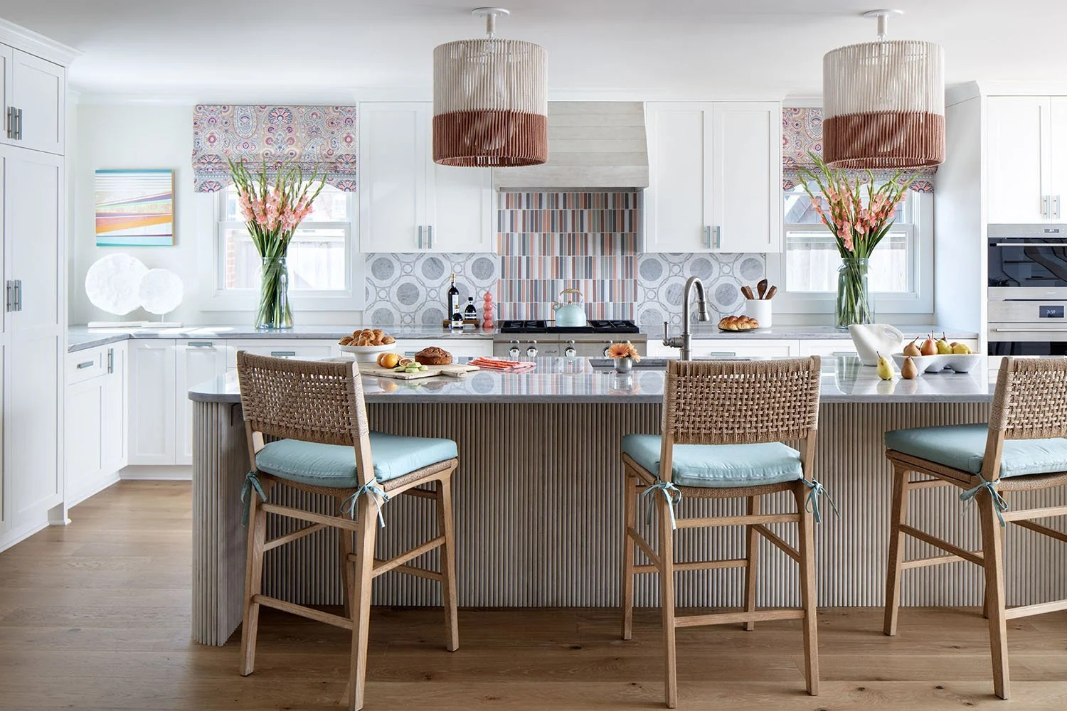 Bright white kitchen with a large island, four wicker bar stools with blue cushions, and colorful backsplash tiles, decorated with pink flowers and breakfast foods.
