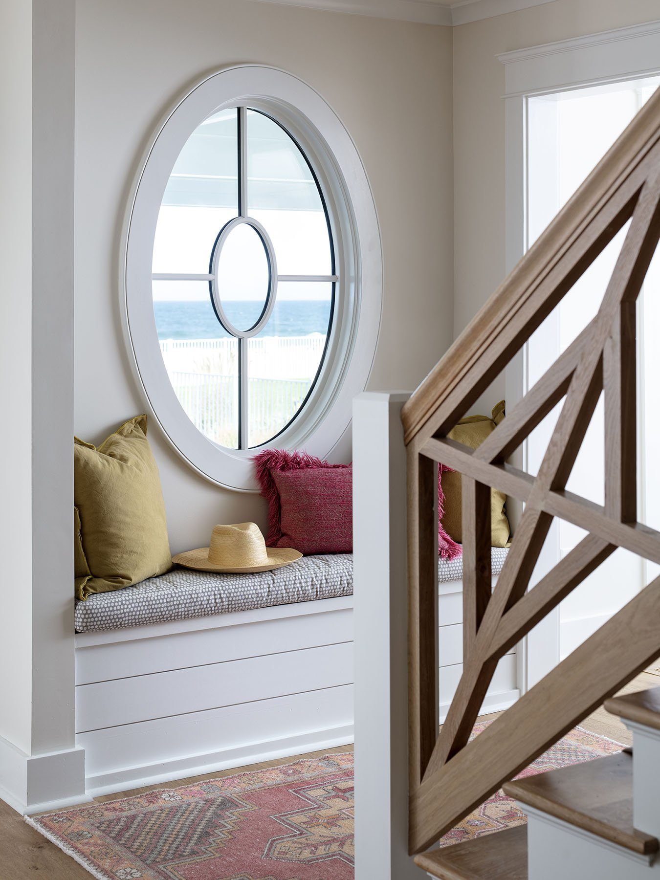 A cozy window nook featuring a built-in bench with pillows and a hat, overlooking the ocean through an oval window in a well-lit home interior.