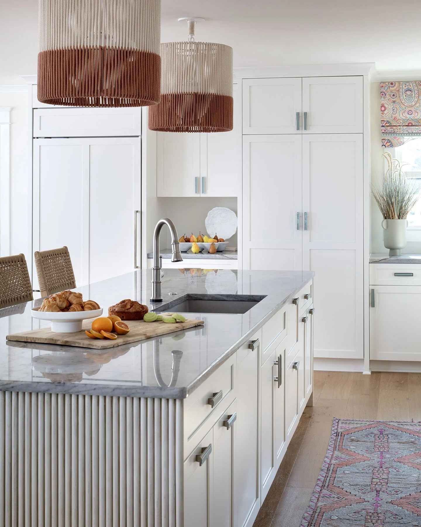 Modern kitchen with white cabinets, marble island counter, stainless steel sink and faucet, hanging pendant lights, fruit bowl, tray of croissants and baked goods, colorful rug, and decorative vase with dried plants.