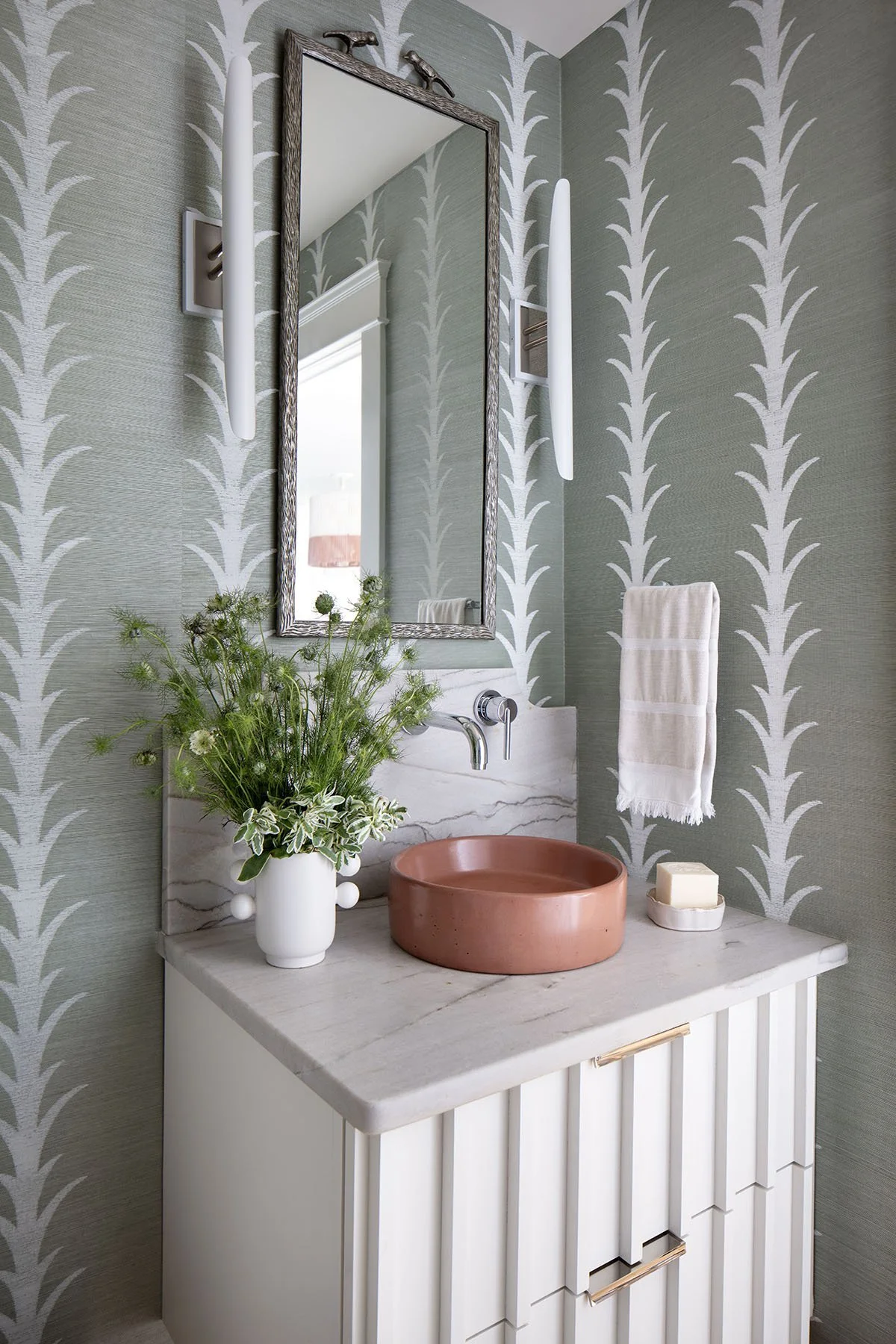 A bathroom vanity with a pink round basin sink, a marble countertop, a mirror with a textured frame, wall-mounted light fixtures, a vase with greenery, a bar of soap, and a towel on the wall.