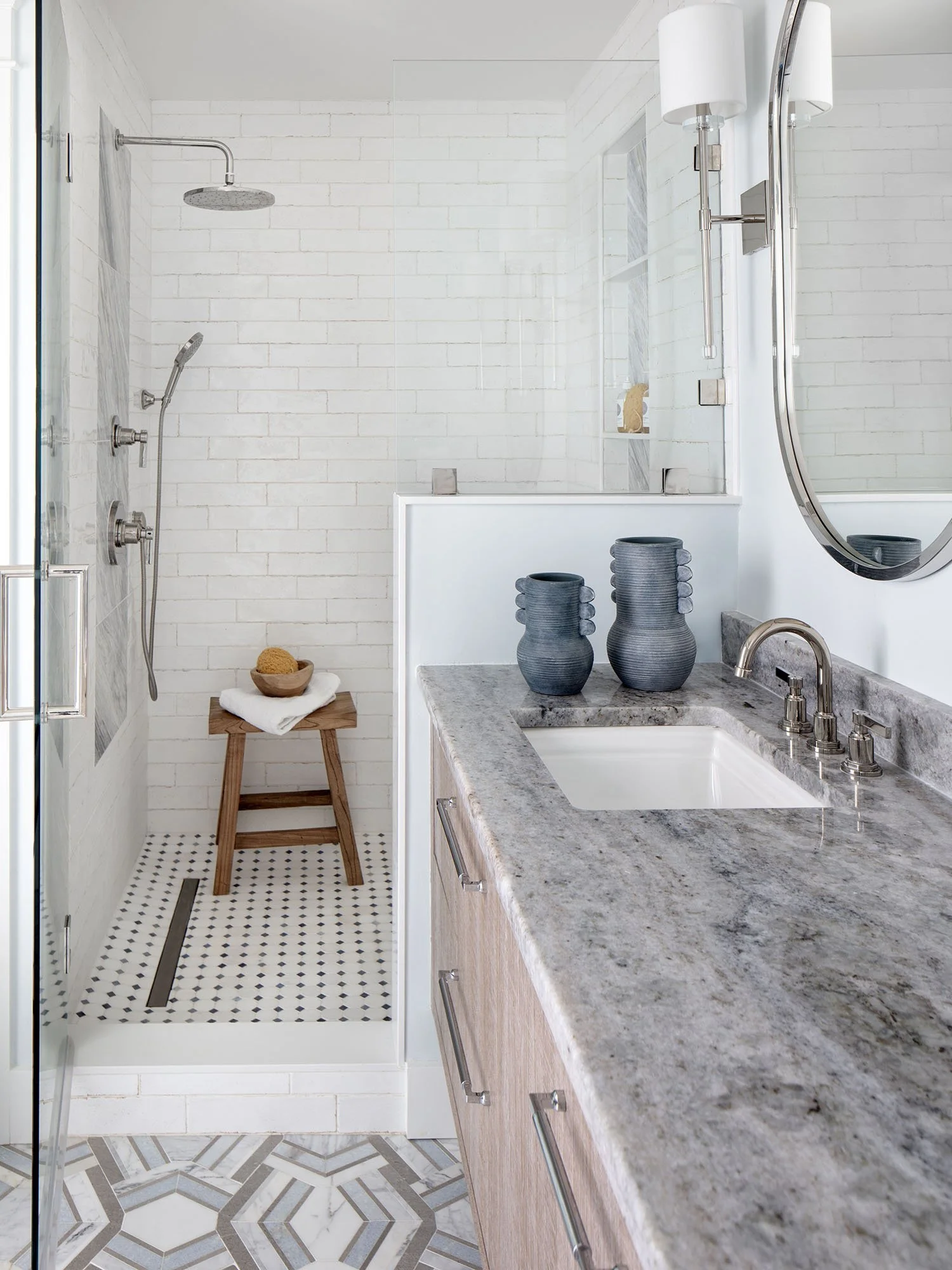 Modern bathroom with a shower area featuring a rain showerhead and handheld shower, light-colored tiled walls, and a vanity with a granite countertop, sink, and decorative vases.