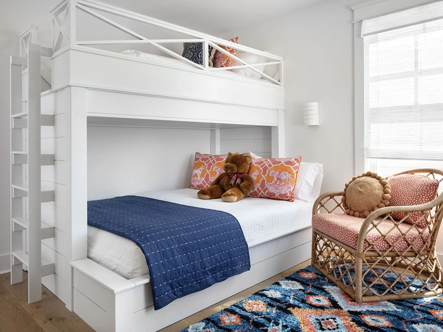A bedroom with a white bunk bed that has a mattress on top and bottom, a brown teddy bear on the bottom bed, pink and orange animal-printed pillows, a woven wicker chair with a crochet pillow, a colorful patterned rug, and a window with white blinds.