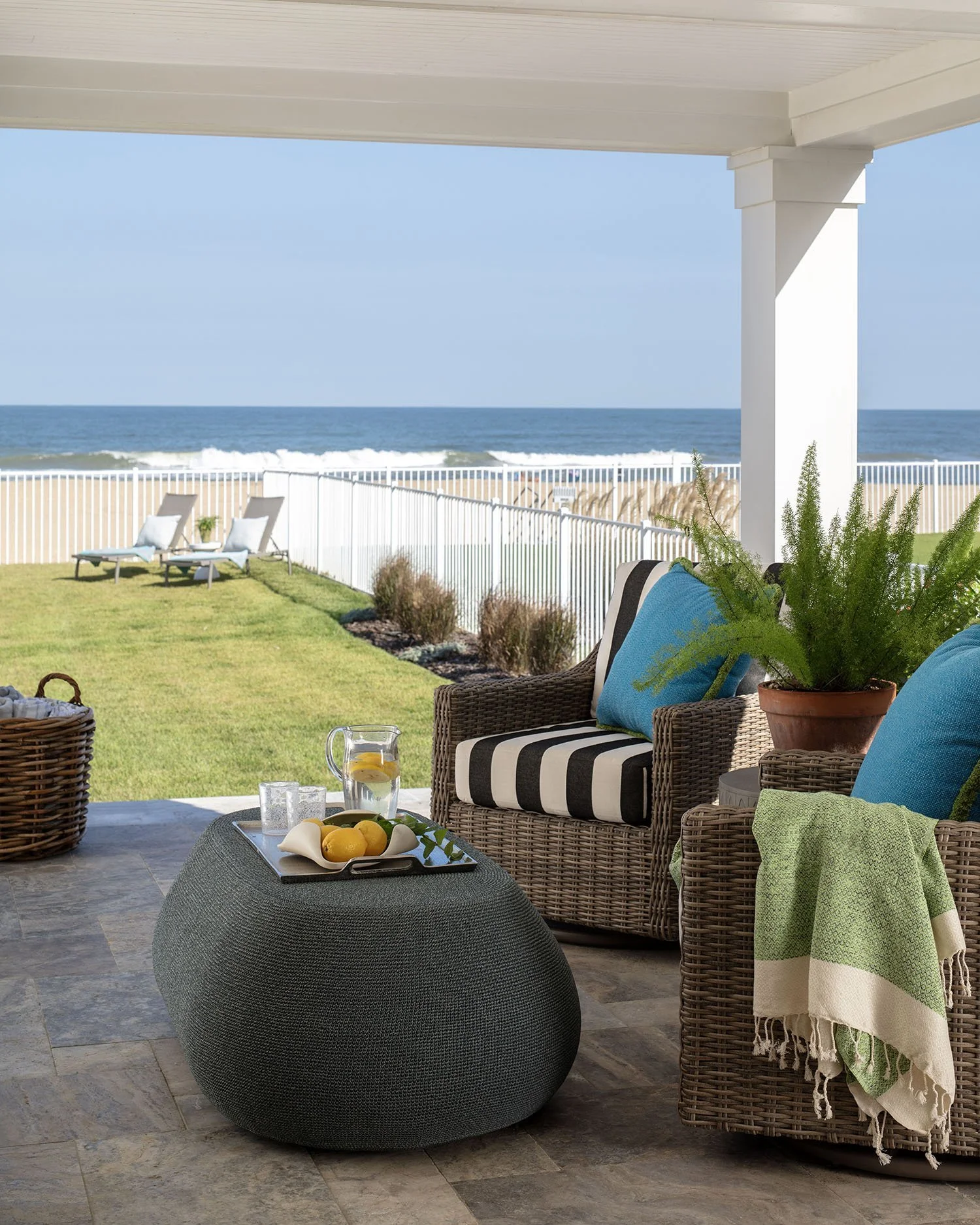 Porch with wicker chairs, blue pillows, green plant, and a table with lemons, pitcher, and drinks overlooking a backyard and ocean view