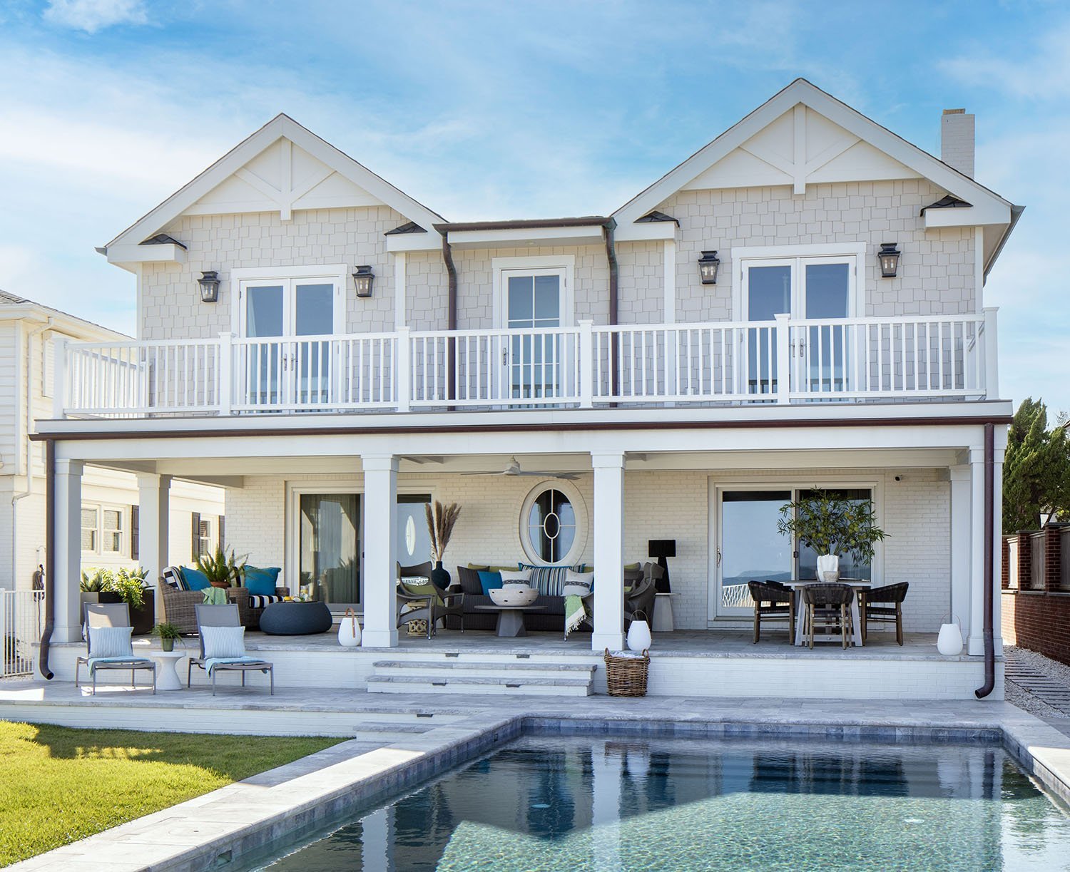 A two-story white house with a large backyard pool, outdoor seating area, and a balcony on the second floor. The house has a gabled roof, glass doors, and windows, with outdoor lighting fixtures.