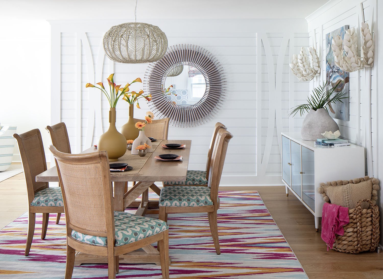 A dining room with a wooden table, surrounded by six chairs with patterned cushions. The table has multiple vases with orange flowers. The room has a white wood-paneled wall with a large round mirror. A white sideboard with decorative items and a lar