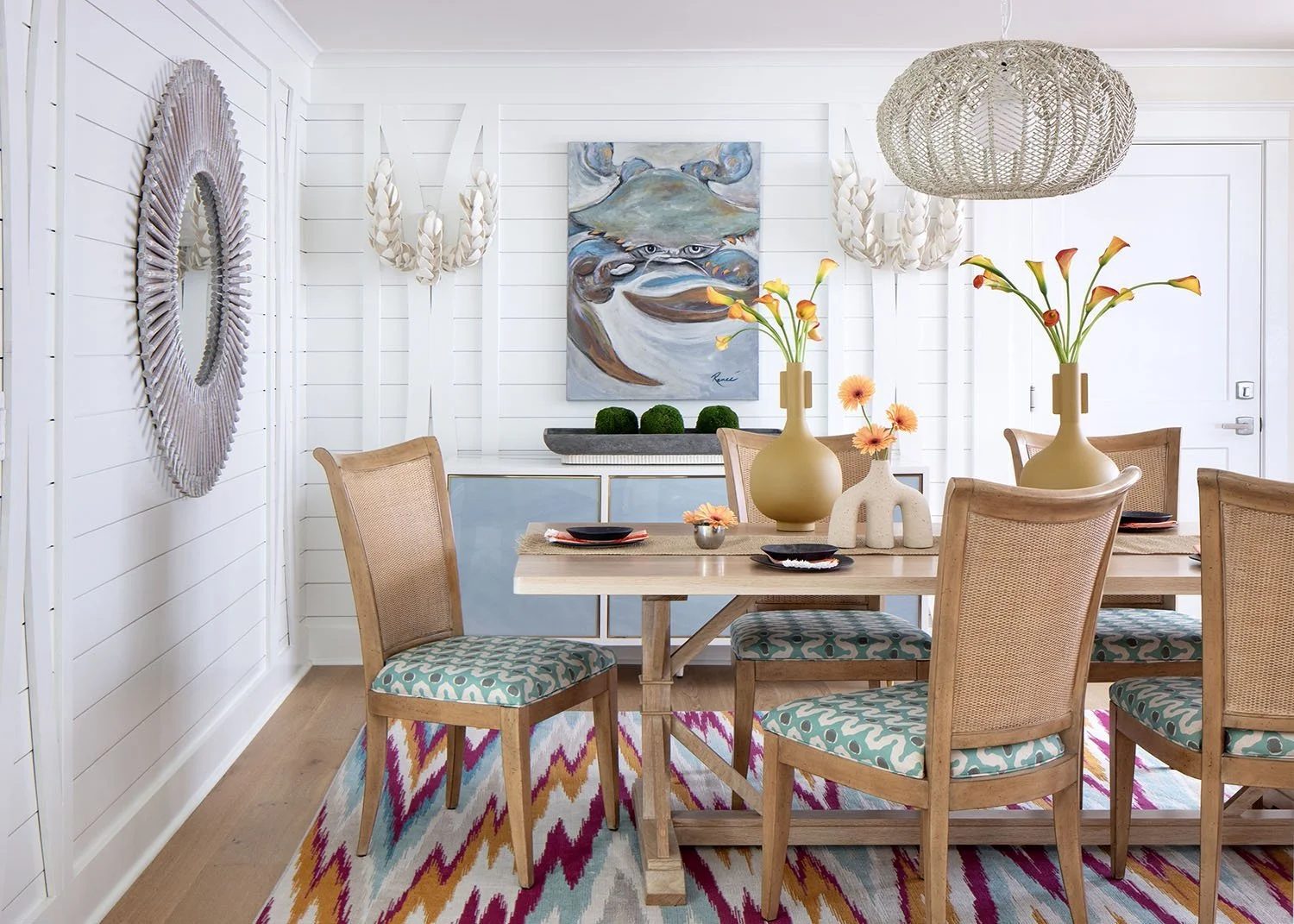 Dining room with white paneled walls, wooden table, patterned chairs, large beige vases with orange and pink flowers, colorful rug, wall art, mirror, and woven pendant light