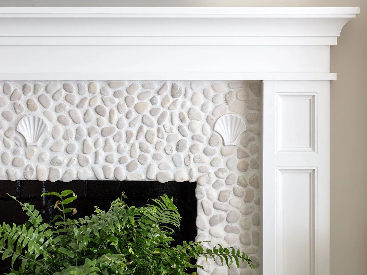 Close-up of a white fireplace with a pebble mosaic and shell decorations, partially covered by green houseplants.