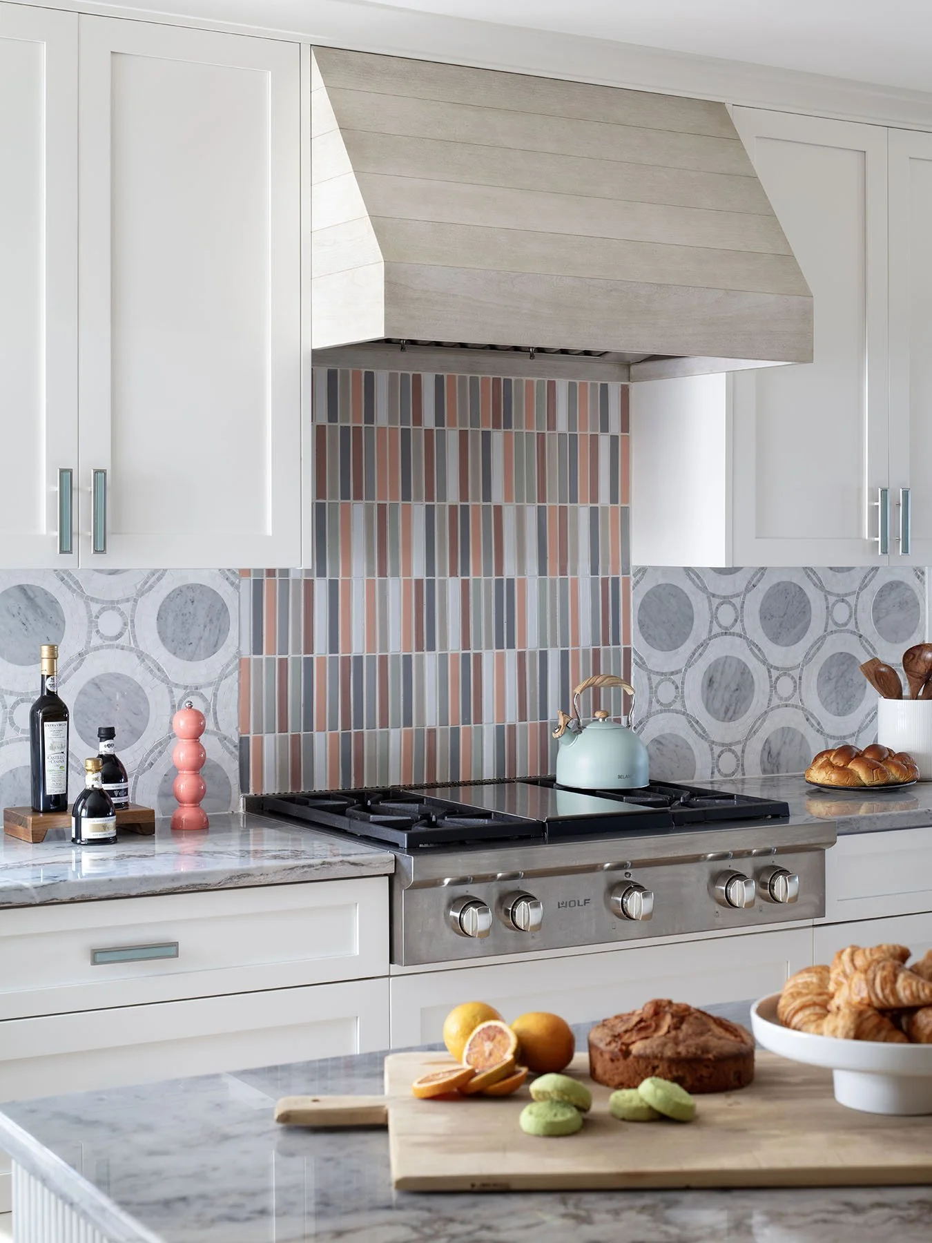 A modern kitchen with white cabinetry, a marble countertop, and a stove with a teal teapot. There are decorative tile backsplashes, a cutting board with fruit and baked goods, and containers with cooking utensils.