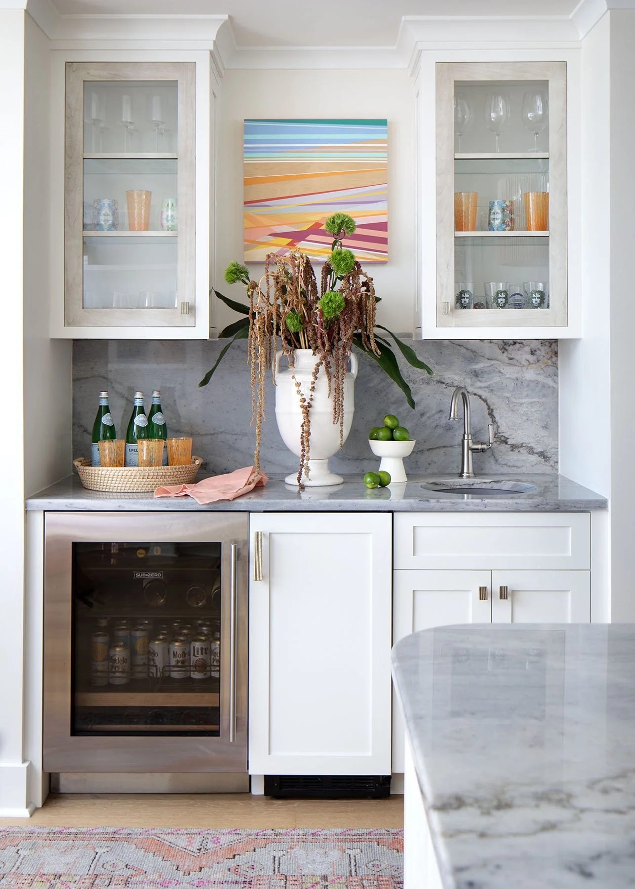 Modern kitchen with white cabinetry, marble countertop and backsplash, glass-front cabinets with glassware, a small sink, a wine fridge, and a decorative vase with green flowers and limes on the counter.