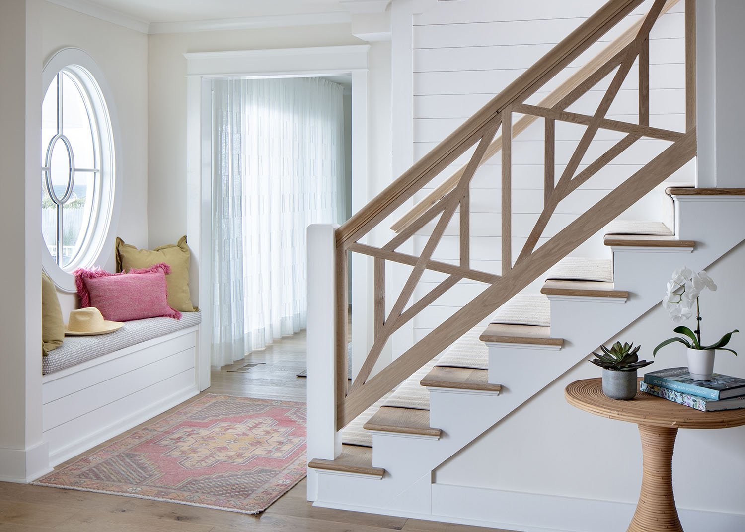Interior view of a cozy home entrance with a built-in window seat with pink and beige cushions, a straw hat, and a light-colored rug; staircase with wooden railing and beige carpeted stairs; a small round table with potted plants and books.