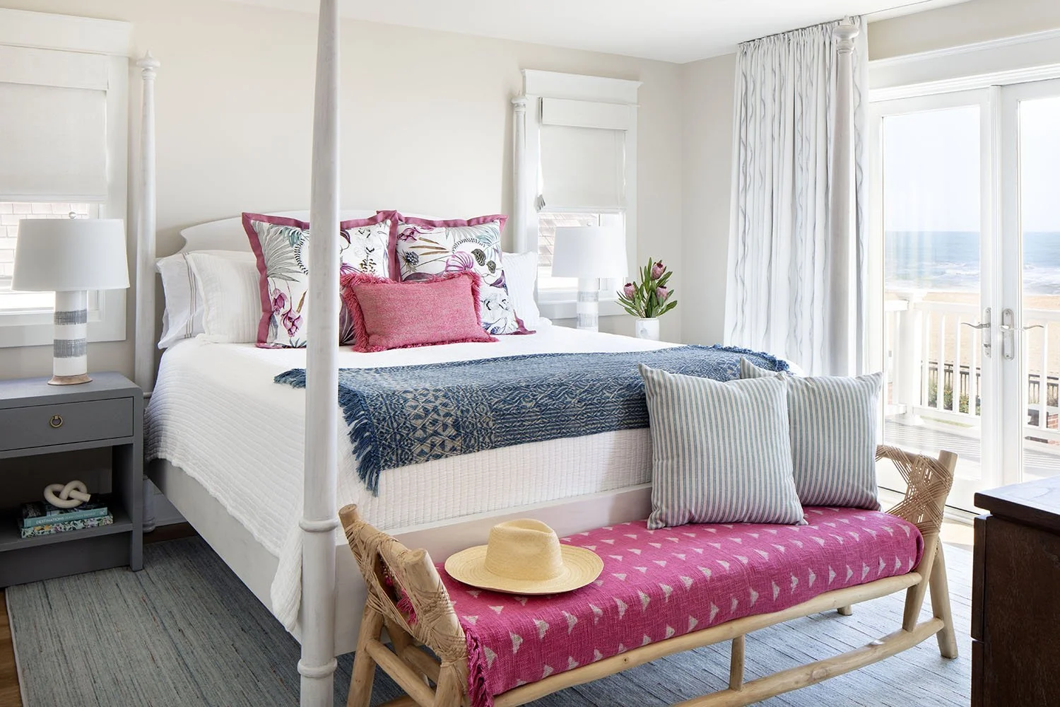 Bright bedroom with white four-poster bed, colorful pillows, blue and pink throws, nightstands with lamps, sliding glass door with ocean view and curtains, wicker bench with pillows and a straw hat inside.