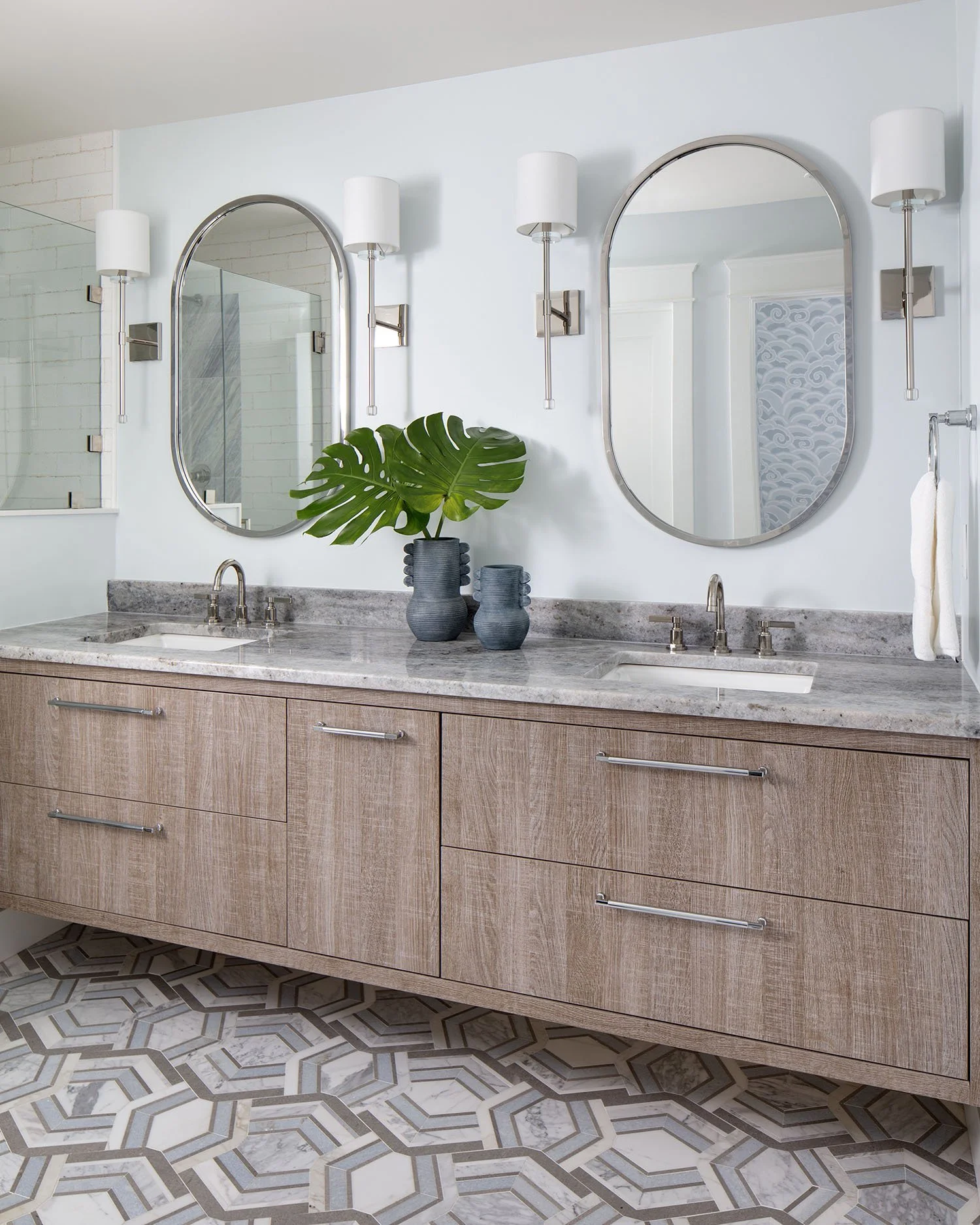 Modern bathroom vanity with a marble countertop, two oval mirrors, wall-mounted sconces, a potted plant, and patterned tile flooring.