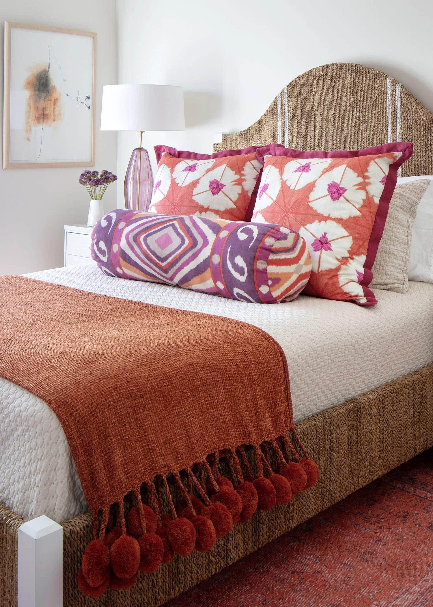 A bed with a woven rattan headboard, white textured bedsheet, orange throw blanket with pom-pom fringe, and decorative pillows including two large pillows with orange and pink leaf patterns, a round bolster pillow with vibrant geometric and floral de