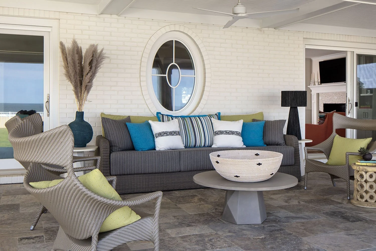 Coastal porch with white brick walls, sliding glass door, black table lamp, sofa with colorful pillows, wicker chairs with green cushions, decorative bowl on round table, and view of ocean through door