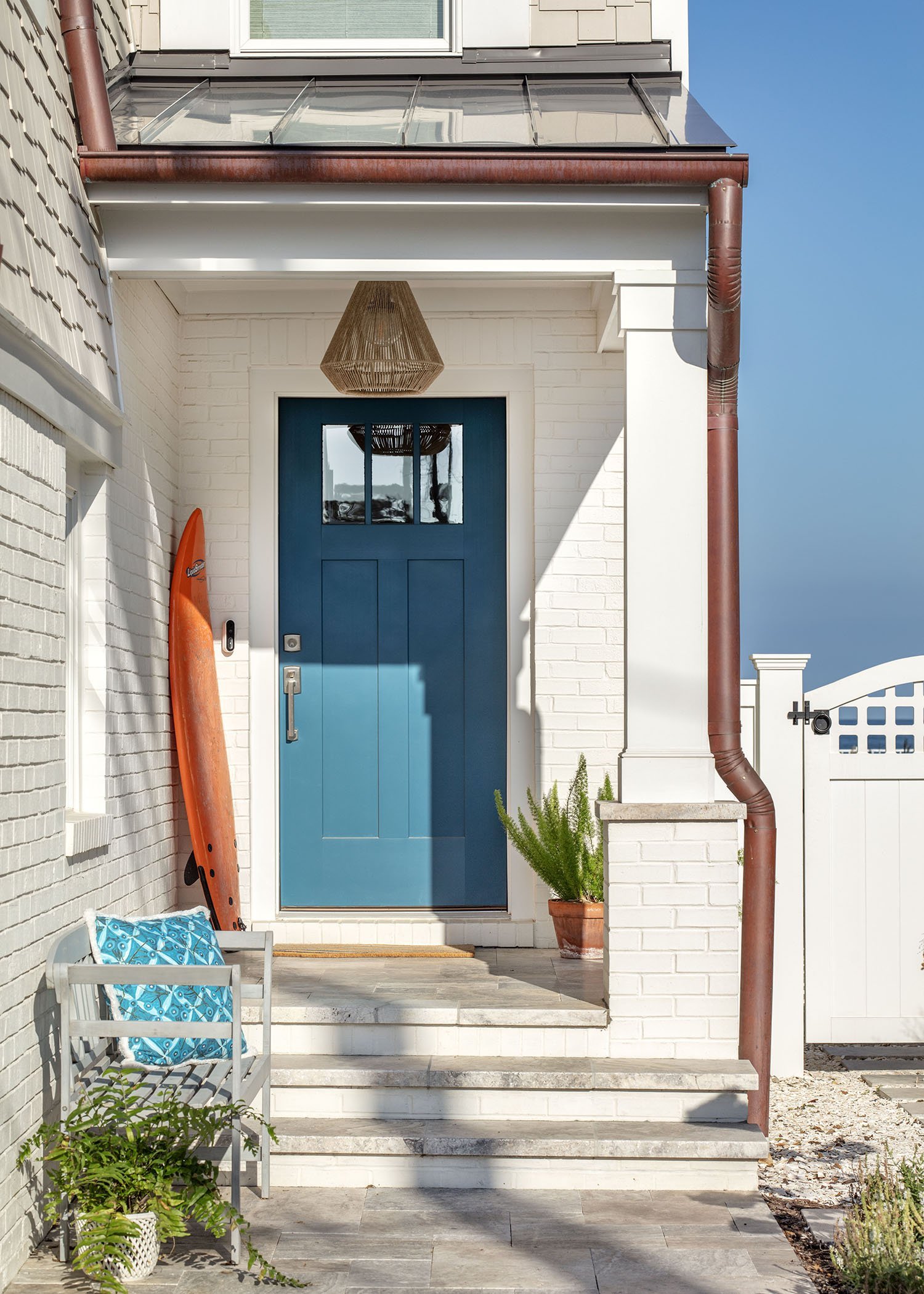 Front porch of a house with a bright blue door, an orange kayak leaning against the wall, a gray bench with a blue cushion, potted plants, and a doormat. The porch has white brick walls and steps, with a brown metal gutter.