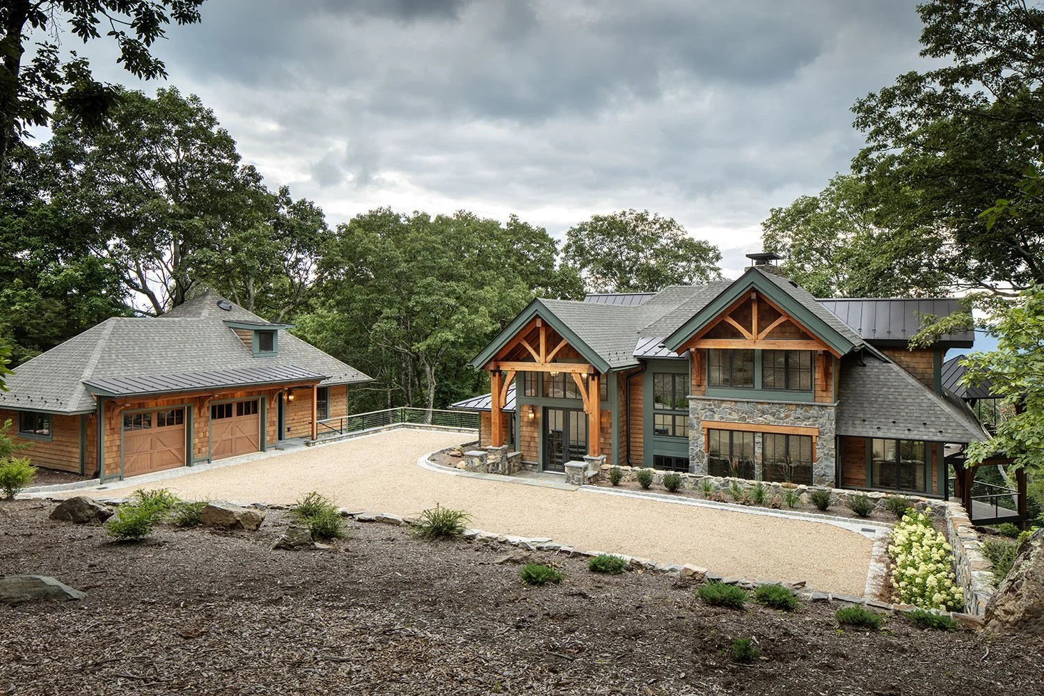A large modern house with a stone and wood exterior, surrounded by trees, under a cloudy sky. There is a spacious gravel driveway and a separate garage with a gabled roof.