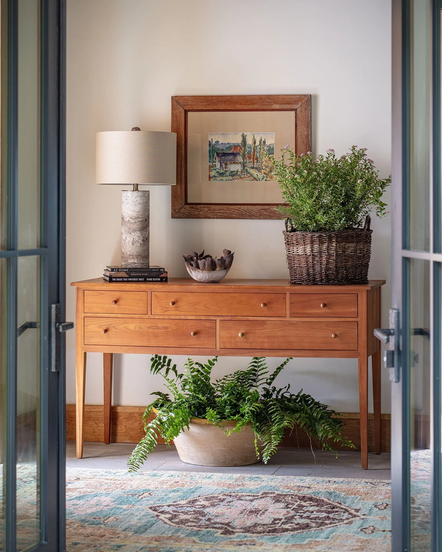 A wooden sideboard with drawers holding a table lamp, a stack of books, a decorative bowl, a potted plant in a woven basket, and a framed painting hanging above. There is a large green fern in a pot on the floor below. The scene is viewed through gla