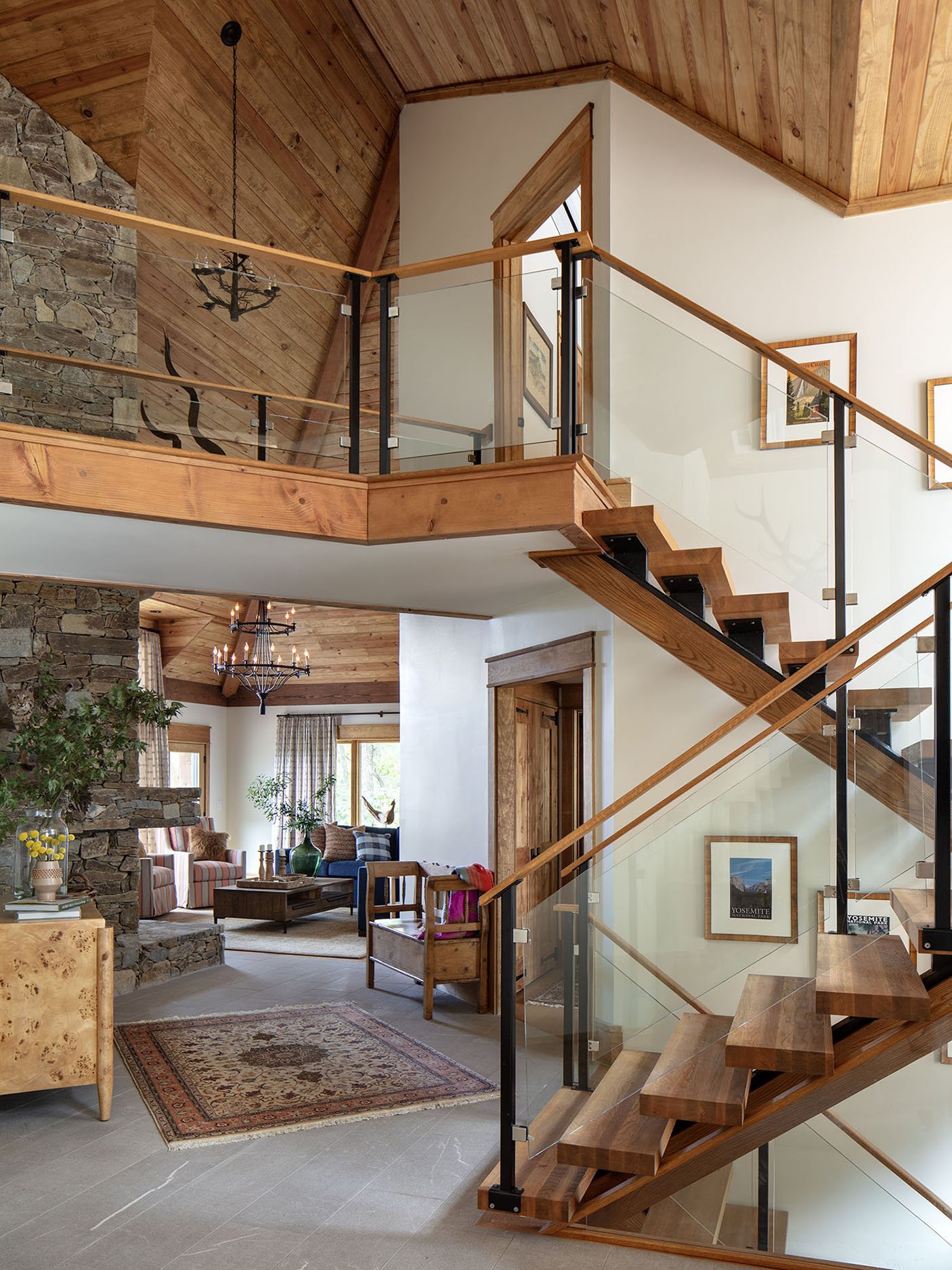 The image shows a modern interior with a wooden staircase and a balcony overlooking a living room with stone fireplace, large windows, and cozy furniture.