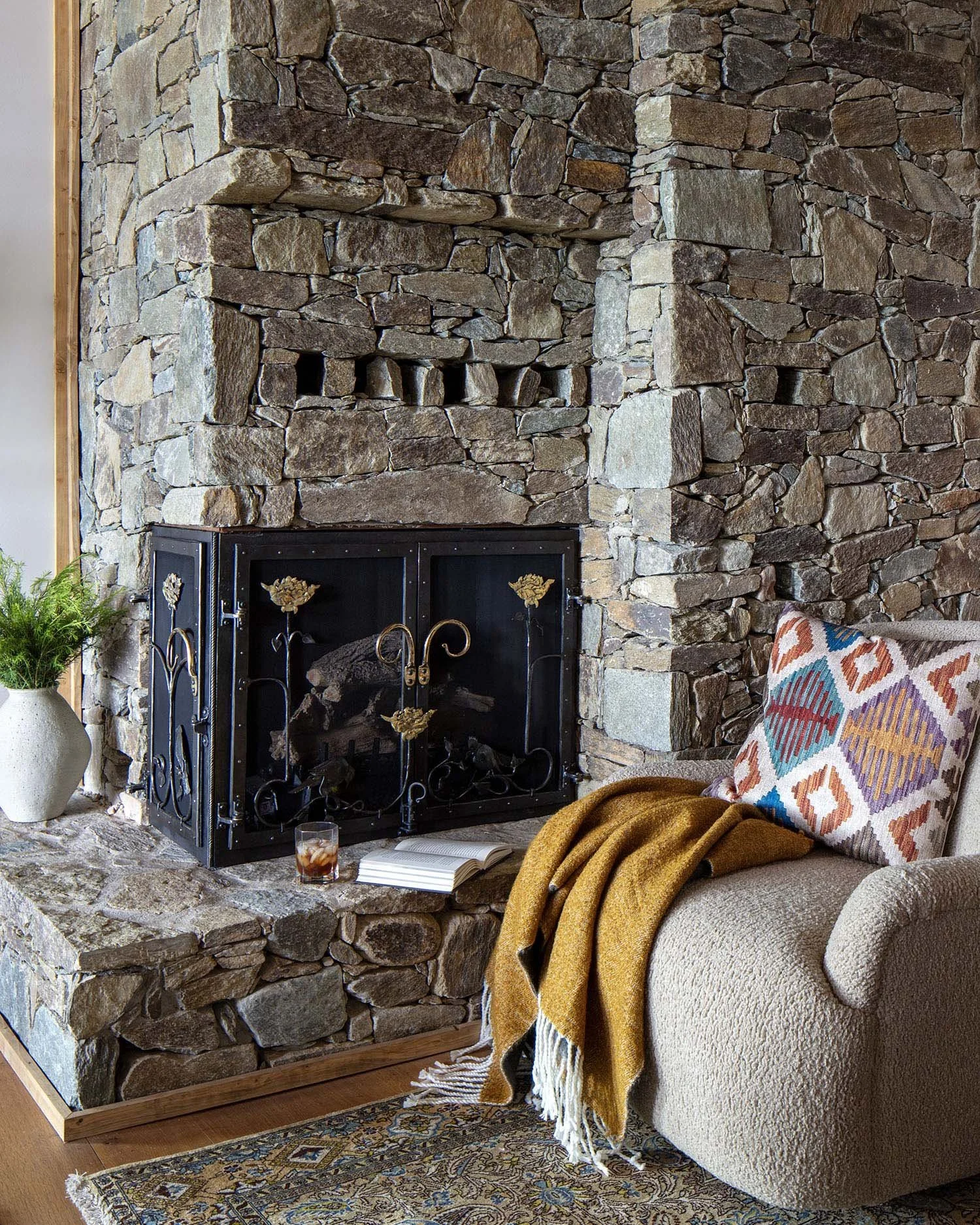 Living room corner with a tall stone fireplace, beige armchair with a colorful patterned pillow, a mustard yellow throw blanket, a book, and a drink on the stone ledge, next to a potted green plant.
