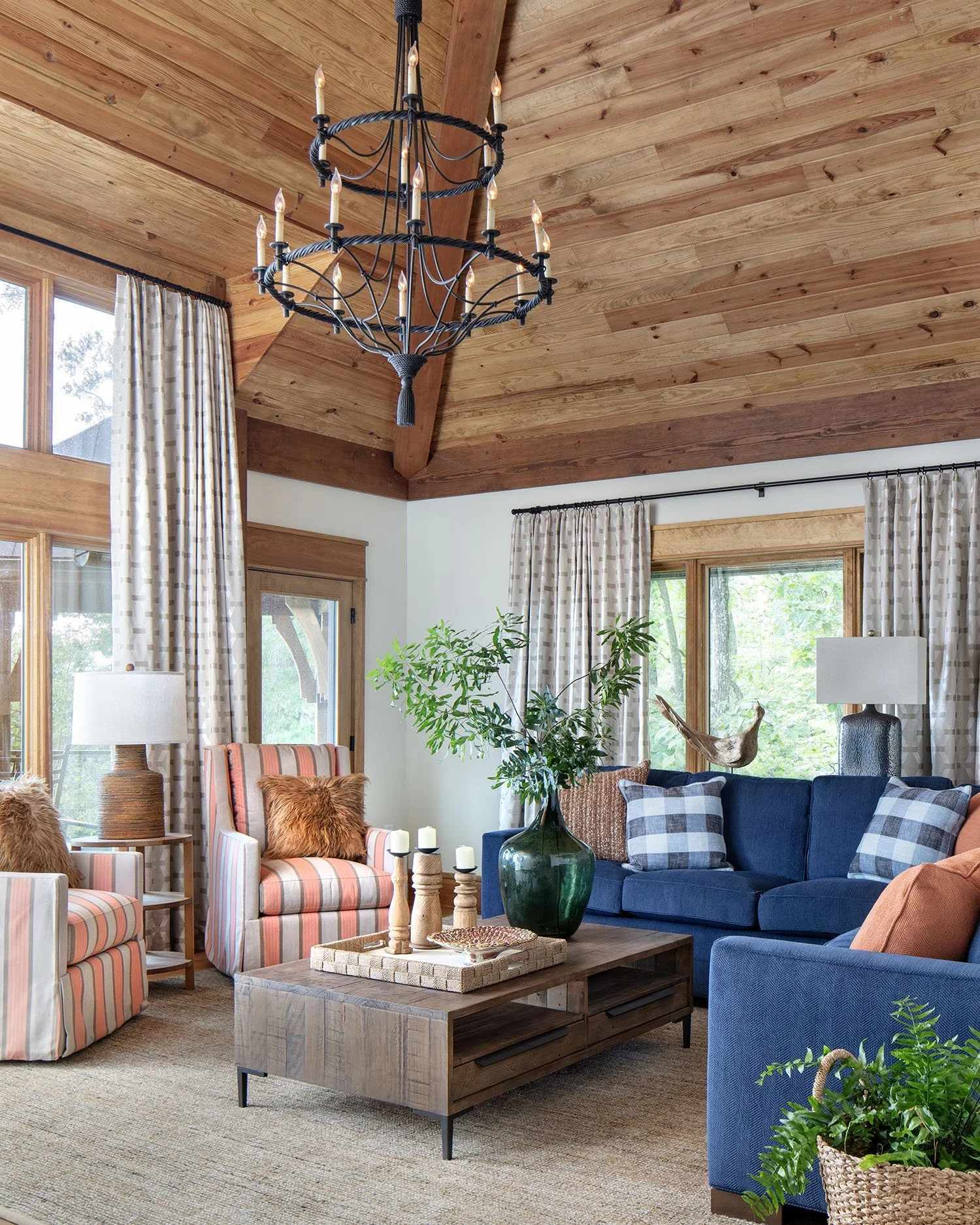Living room with wood-paneled vaulted ceiling, large windows with curtains, a large blue sofa, striped armchair, wooden coffee table, and decorative elements like plants, candles, and lamps.
