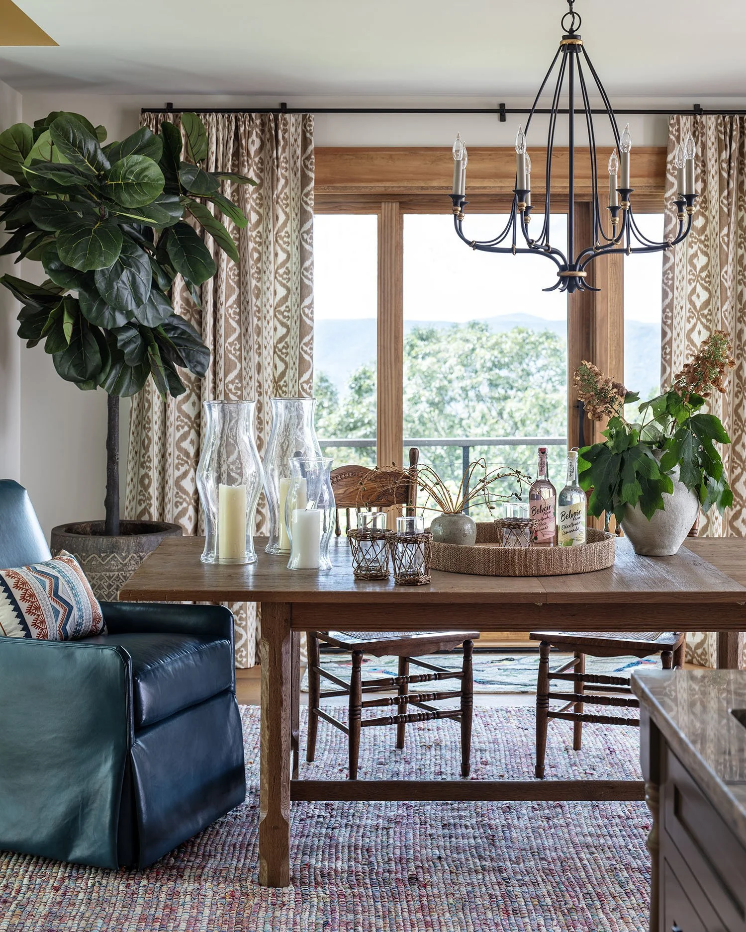 A dining room with large windows showing an outdoor view of trees and hills. The room has a wooden dining table decorated with candles, glass bottles, and potted plants. A black chandelier hangs from the ceiling. There are patterned curtains and a la