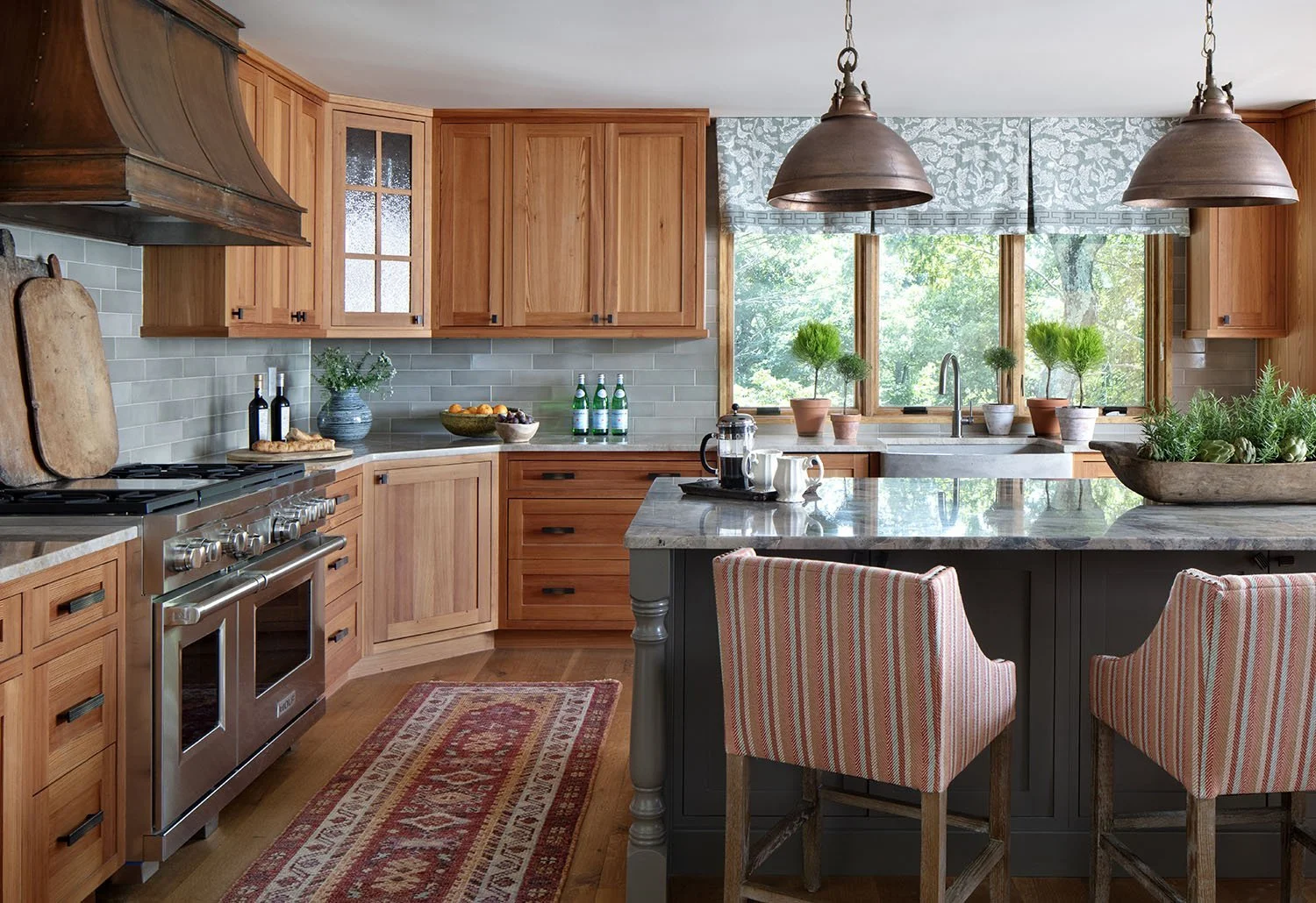 A rustic kitchen with wooden cabinets, a kitchen island with a granite countertop, and large windows behind the sink showing greenery outside. Hanging above are two large pendant lights, and the countertop has small potted plants and kitchenware.