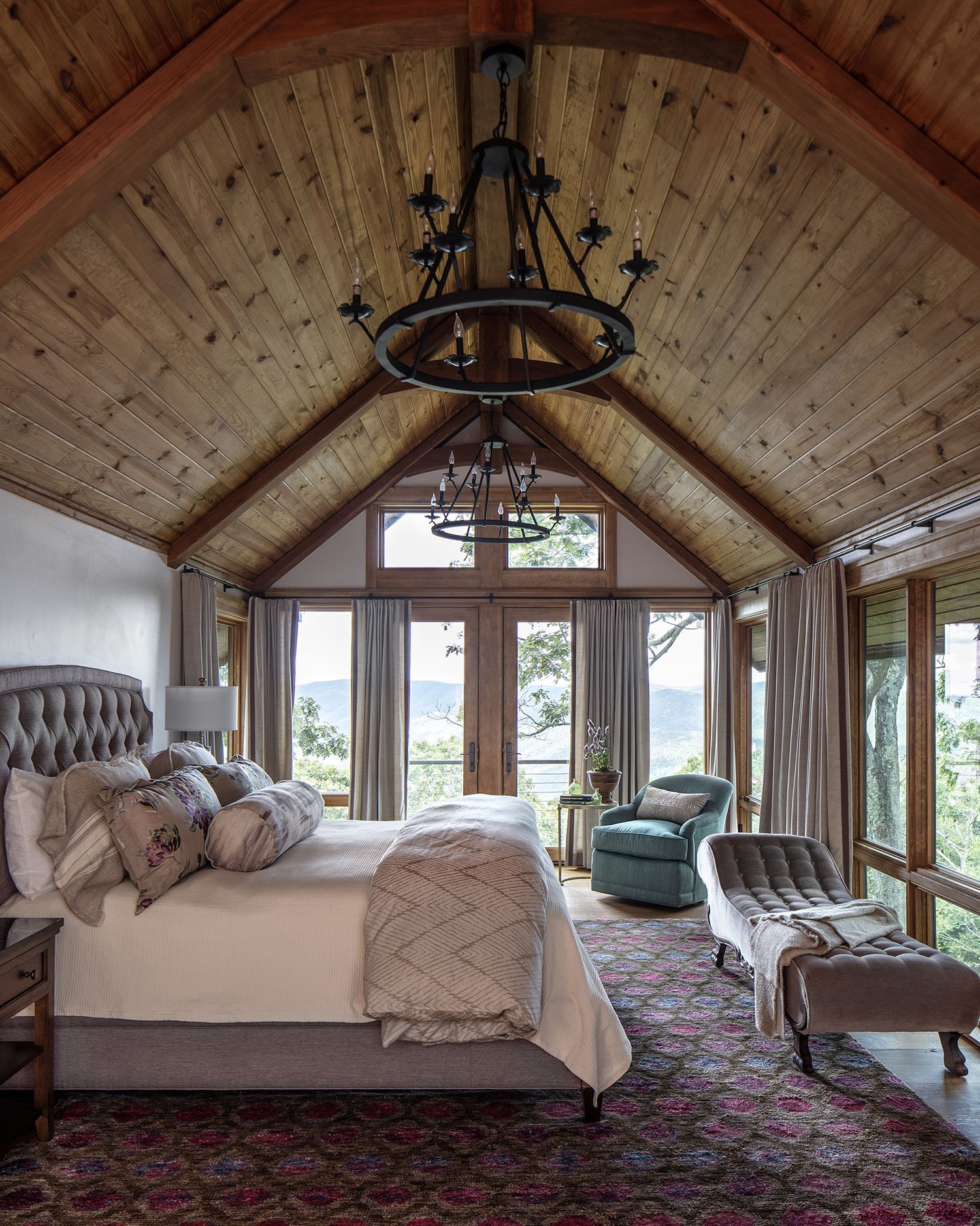 A cozy bedroom with a vaulted wood ceiling, featuring two black chandeliers. The room has large windows with beige curtains, offering a view of trees and mountains. There is a large bed with a tufted headboard, decorated with multiple pillows and a b