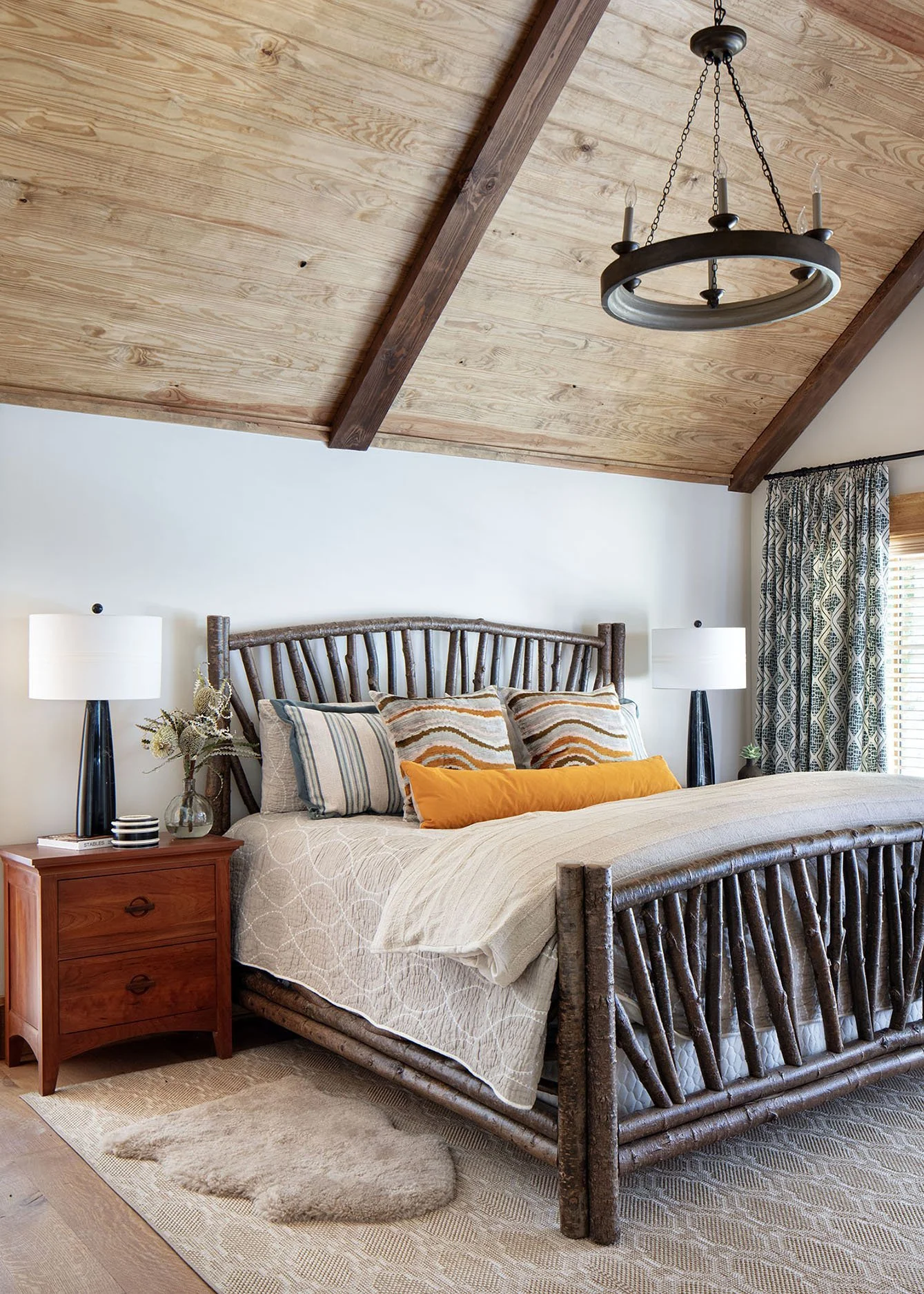 A bedroom with a rustic wooden bed frame, beige bedding, decorative pillows, two nightstands with lamps, a patterned curtain, a wood ceiling with beams, a chandelier, a small plant, and a sheepskin rug.