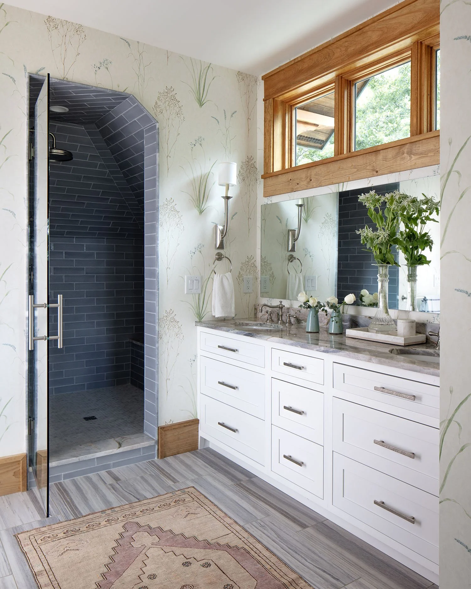 Bathroom with a walk-in shower with dark gray tile, a white vanity with marble countertop, a large mirror, two wall-mounted lights, and a window with wooden trim. Decor includes vases with flowers and a patterned rug on the floor.