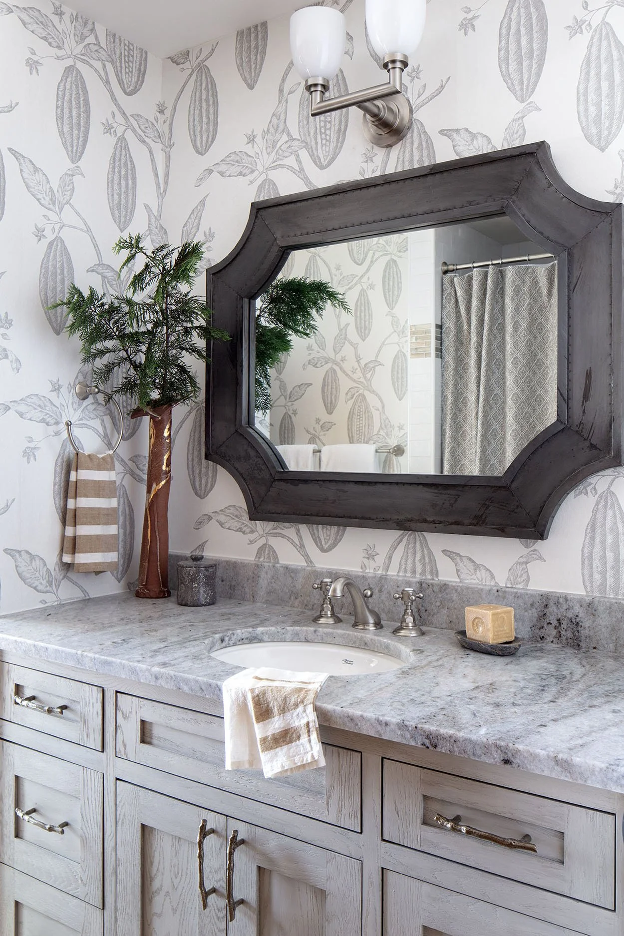 A bathroom vanity with a marble countertop, a sink with a silver faucet, a large dark wooden framed mirror, a wall-mounted light fixture, a small potted plant, a soapdish with soap, a towel hanging from a hook, and a striped towel on the counter, wit