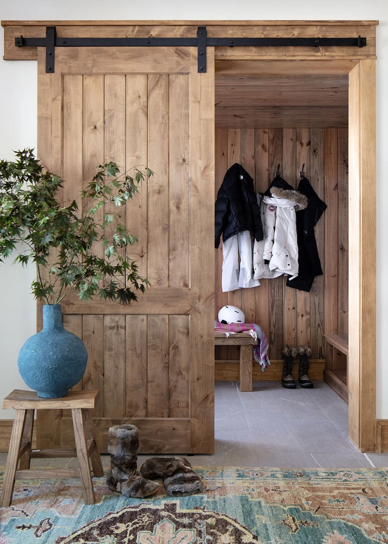 A wooden mudroom with hooks holding winter jackets and boots, a bench, a helmet, a scarf, a pair of boots, a blue vase with a green plant on a small wooden table, and a multicolored rug on the floor.