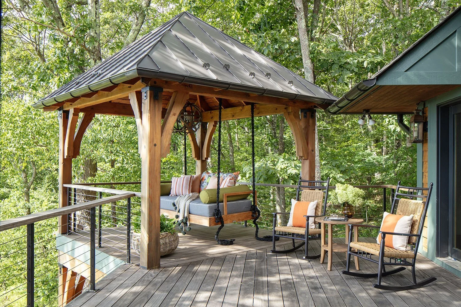 An outdoor wooden deck with a swing, two rocking chairs, and a small table, surrounded by lush green trees.