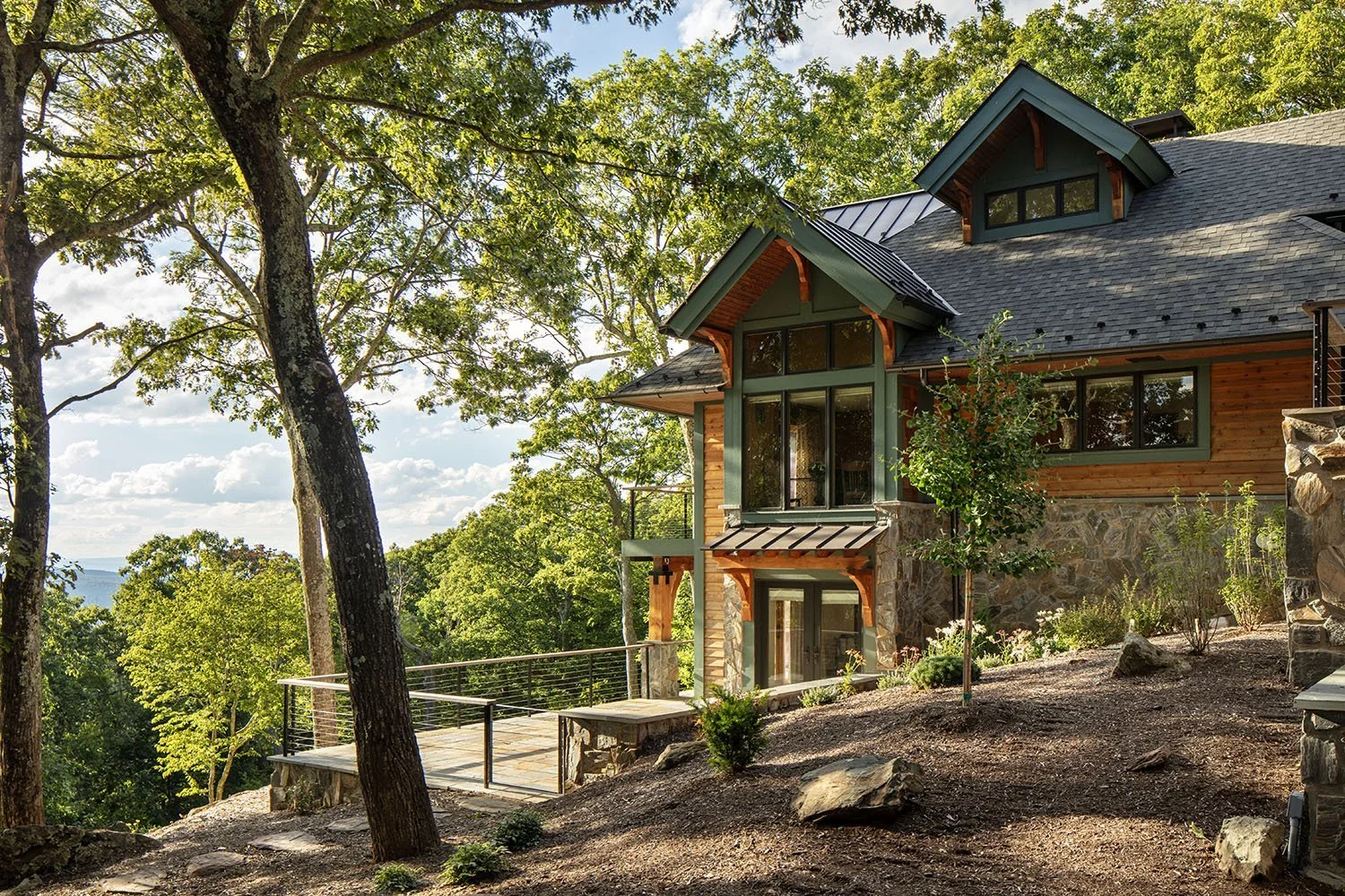 A modern house with large glass windows and a stone foundation is situated on a hillside surrounded by green trees. There is a wooden deck with a metal railing extending from the house, overlooking a wooded landscape. The sky is partly cloudy with su