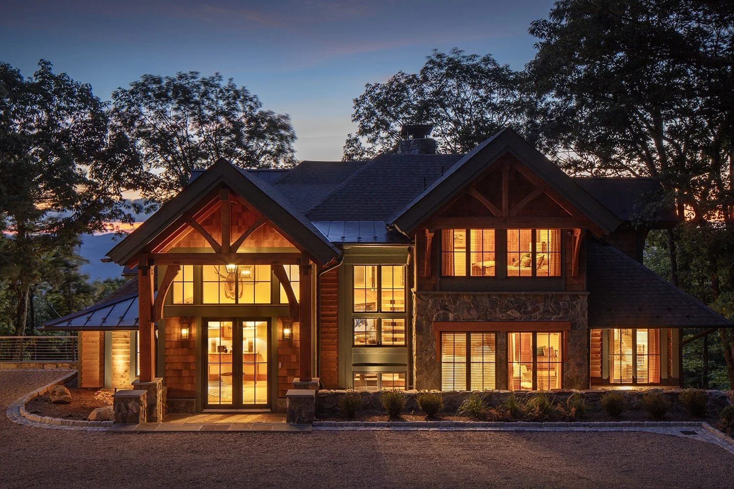 A large house with warm lit windows at dusk, surrounded by trees and a gravel driveway in the foreground.