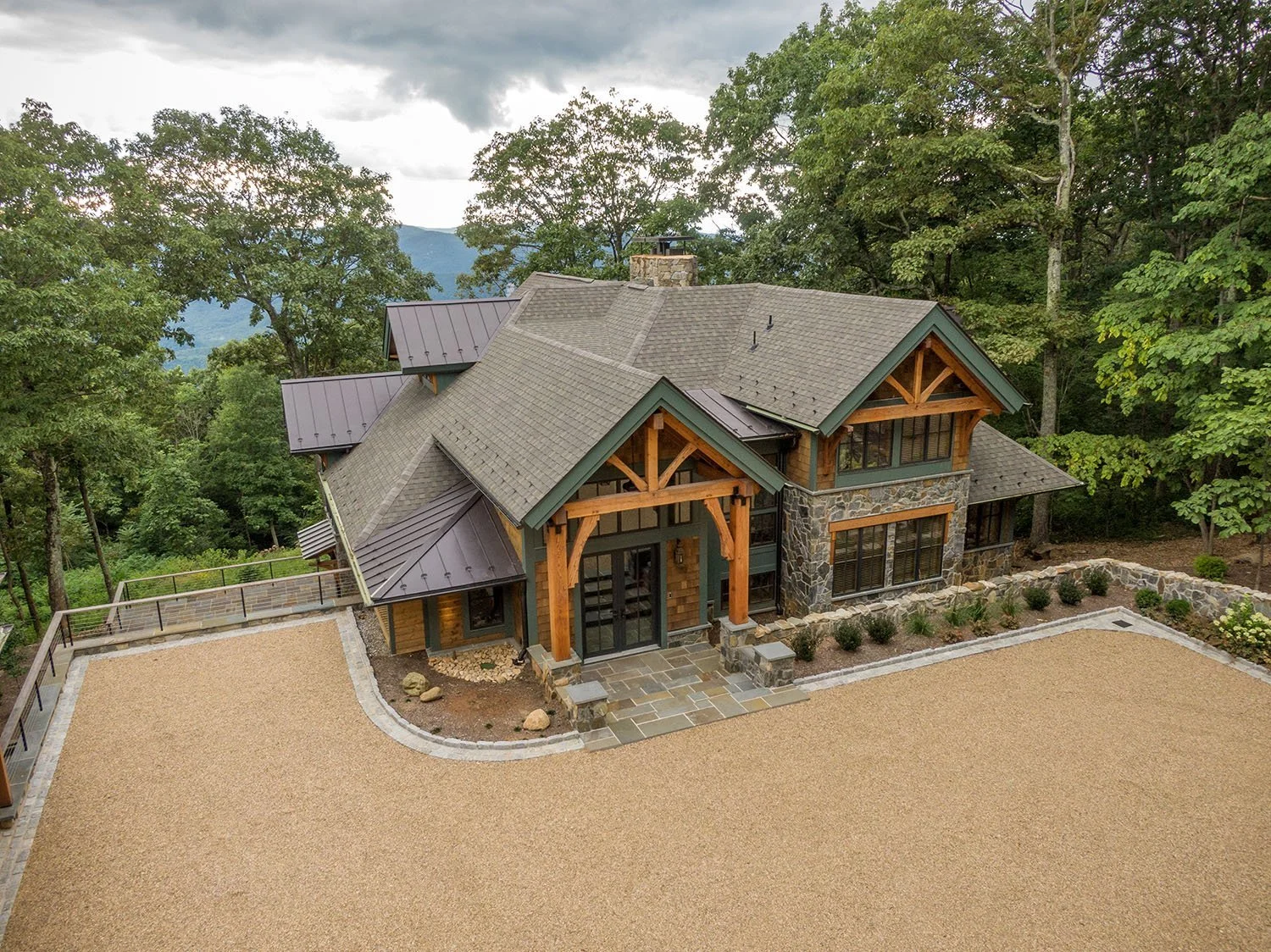 A large house with a stone and wood exterior, multiple gabled roofs, surrounded by trees, and a gravel yard.