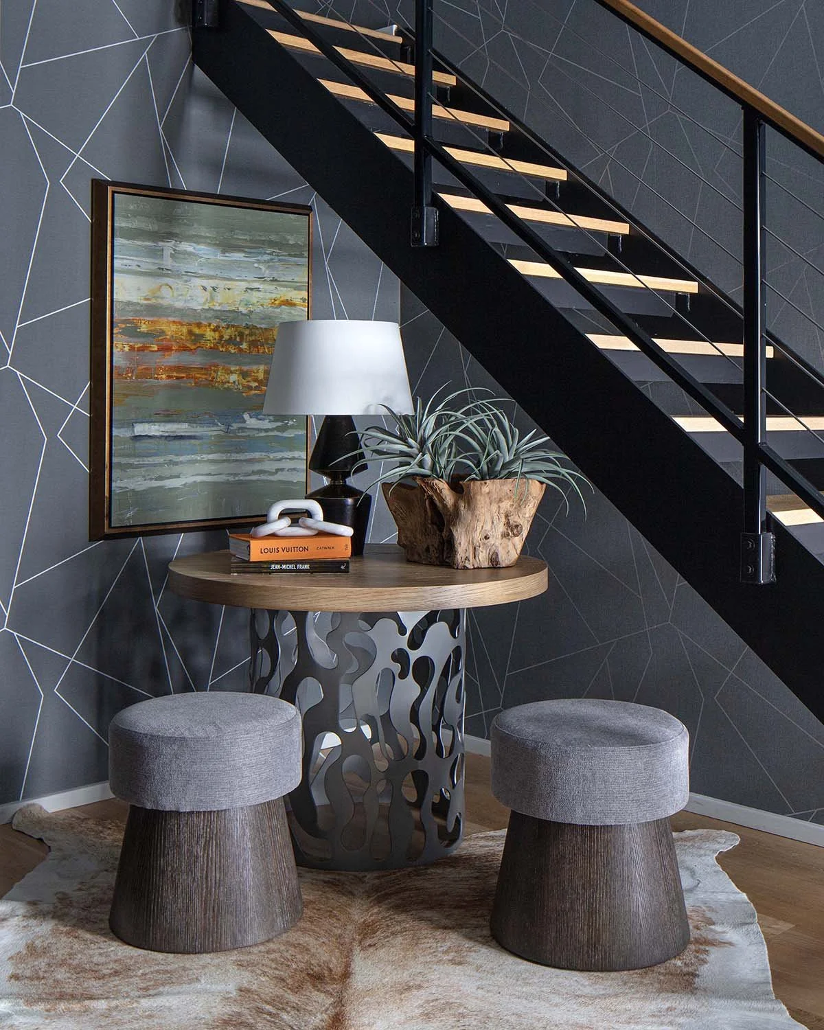 A modern interior with a round wooden table under a staircase, decorated with a large potted plant, a black lamp and books, against a dark geometric patterned wall.