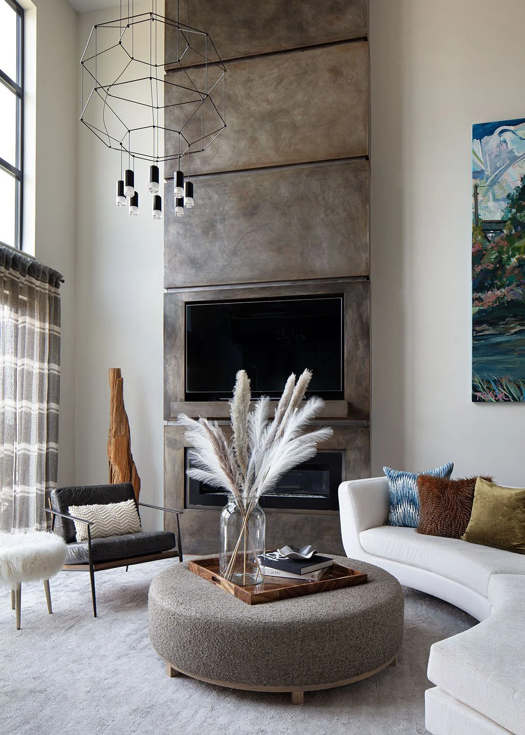 Modern living room with a high ceiling, a fireplace, a mounted flat-screen TV, and contemporary furniture including a white sofa with colorful pillows, a black armchair with a pillow, a round beige ottoman with a tray and a large glass vase with pamp