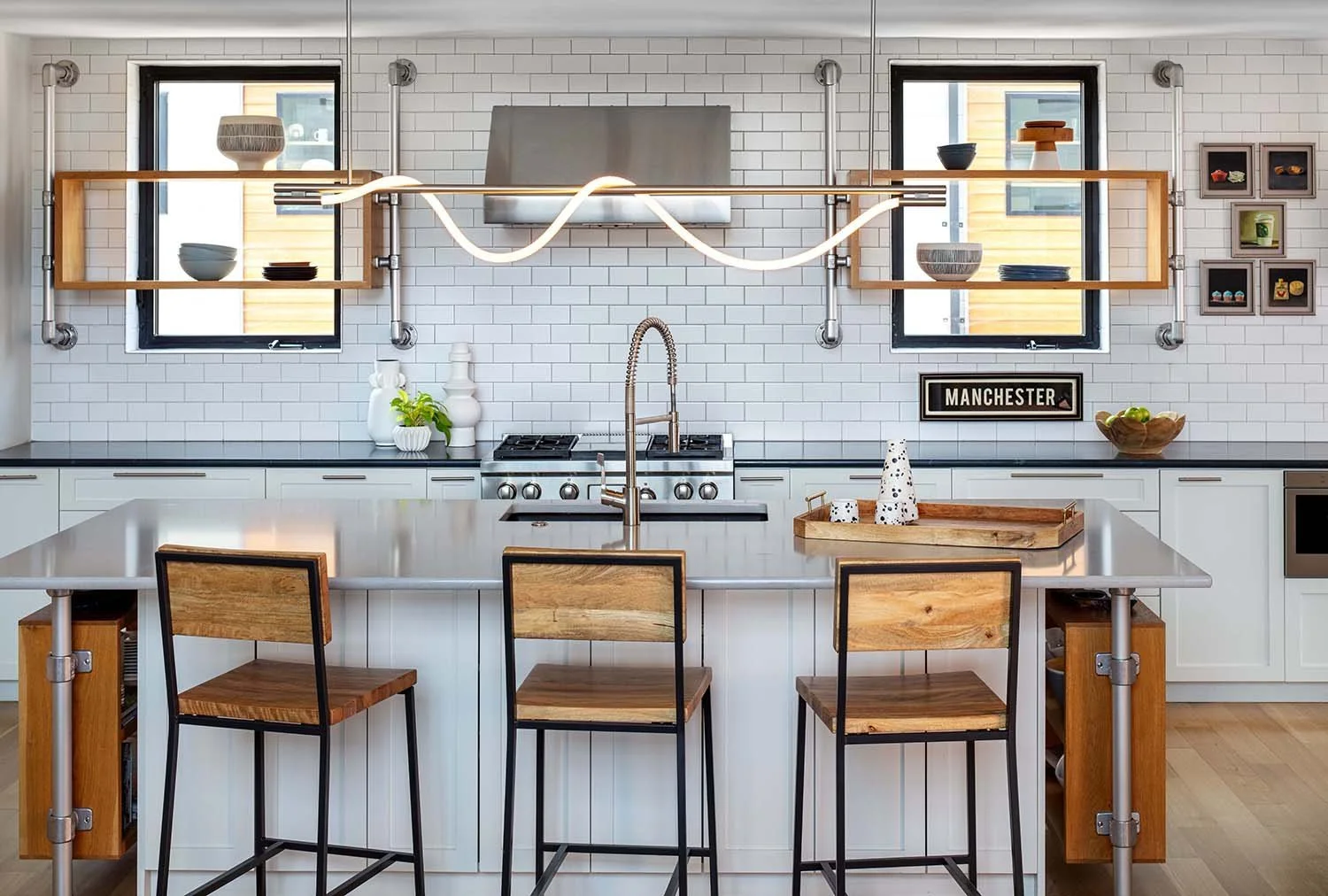 Modern kitchen with white subway tile backsplash, black countertops, and wooden shelving. There are three bar stools with wooden seats and black metal frames at a white kitchen island. The kitchen has two small black-framed windows, a wall-mounted TV