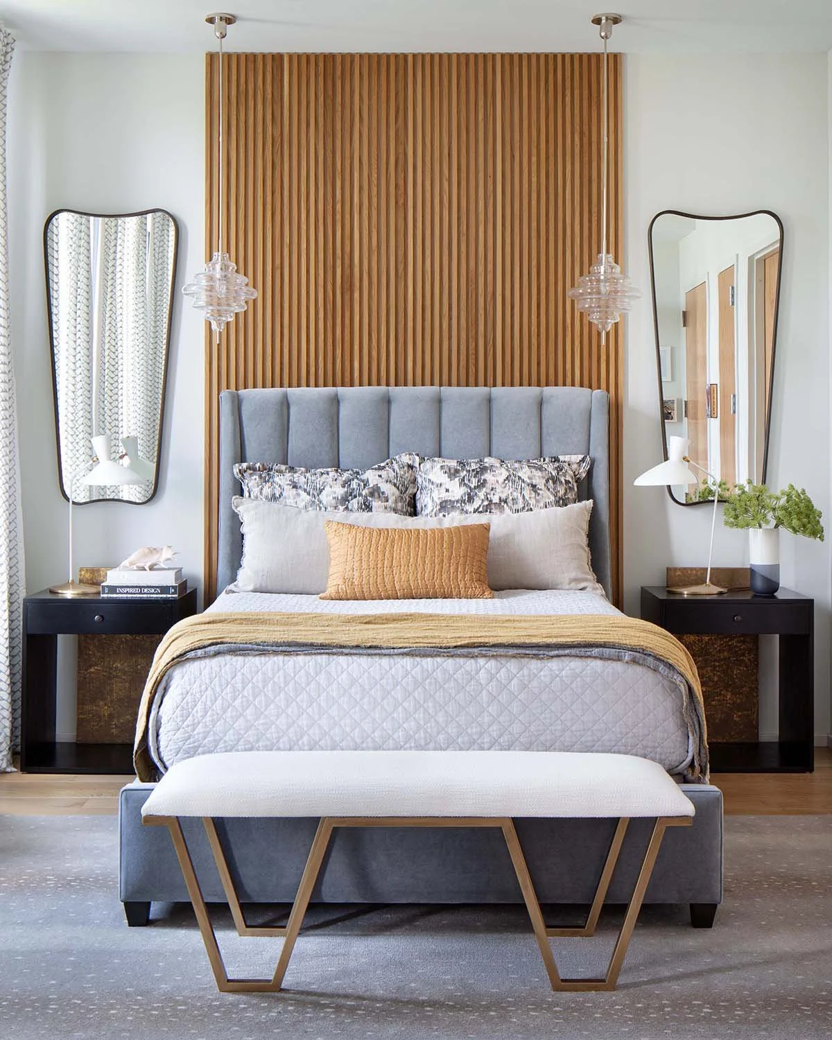 A modern bedroom with a gray upholstered bed, decorative pillows, and a beige quilt. Two black nightstands with lamps and books are on each side of the bed. Behind the headboard is a wooden slat accent wall, with two hanging pendant lights. Two leani