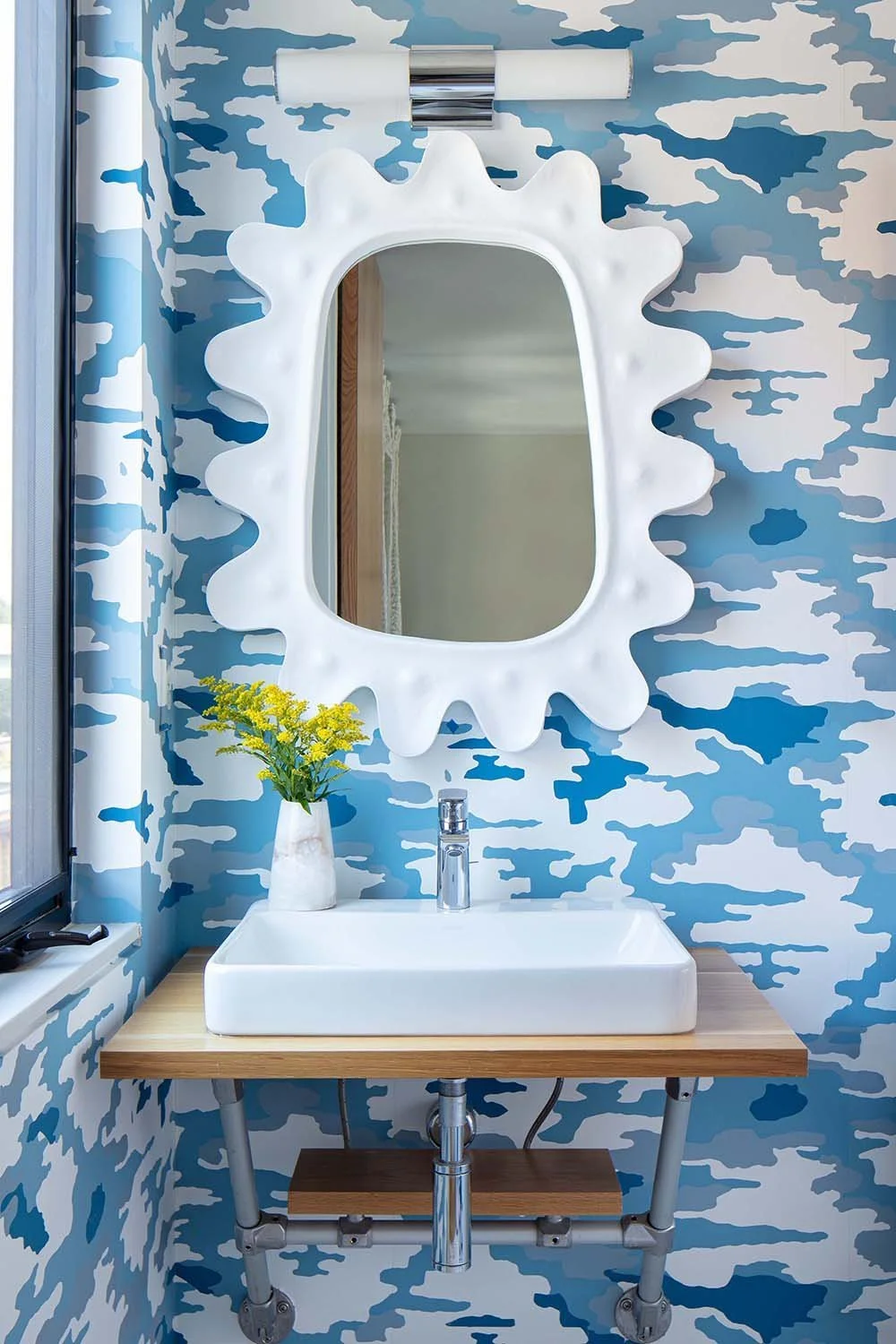 Modern bathroom with a blue camouflage-patterned wallpaper, a white soap basin, a small wooden shelf, a tall white vase with yellow flowers, a silver faucet, and a uniquely shaped white mirror.