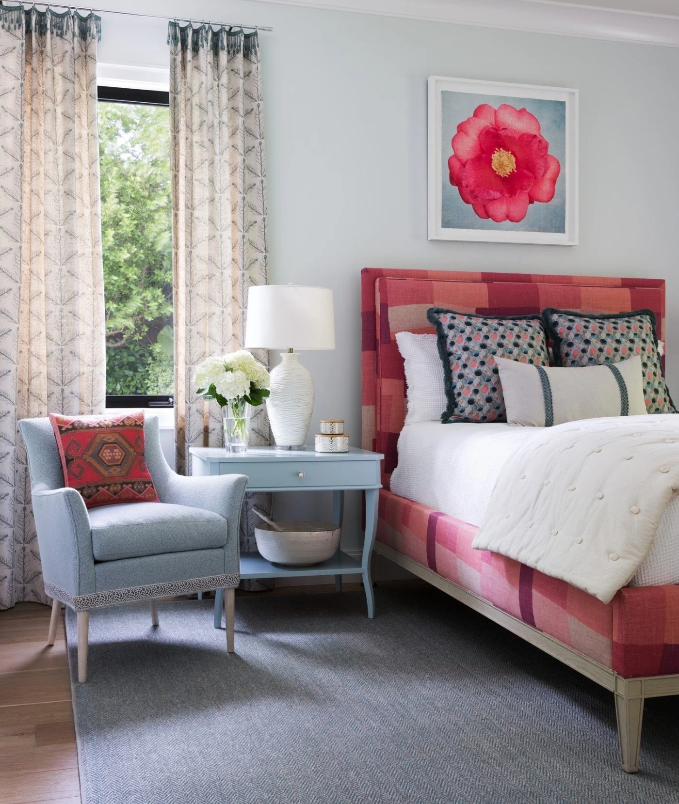 A cozy bedroom featuring a bed with a pink checked headboard and white bedding, a light blue nightstand with a white lamp and floral arrangement, a light blue armchair with a patterned pillow, and framed floral artwork on the wall, with window curtai