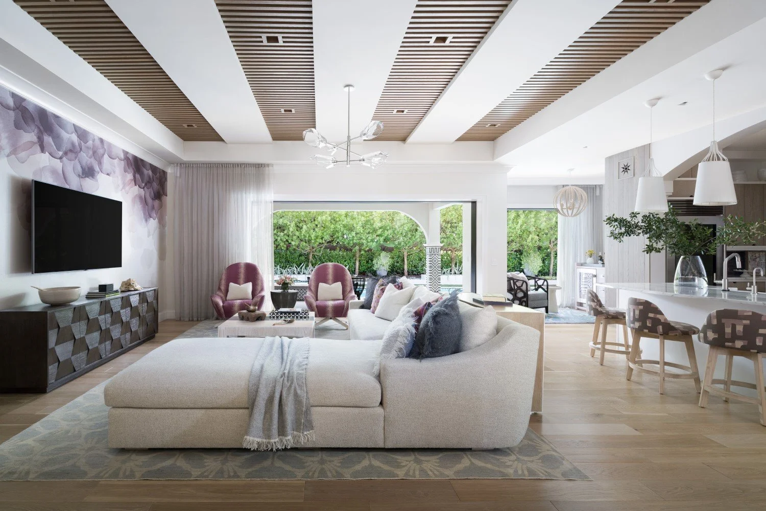 Modern living room with large beige sectional sofa, pink armchairs, and a television on a textured black media console. Open kitchen with white island, bar stools, and greenery in a glass vase. Large window showing trees outside, light wood flooring,