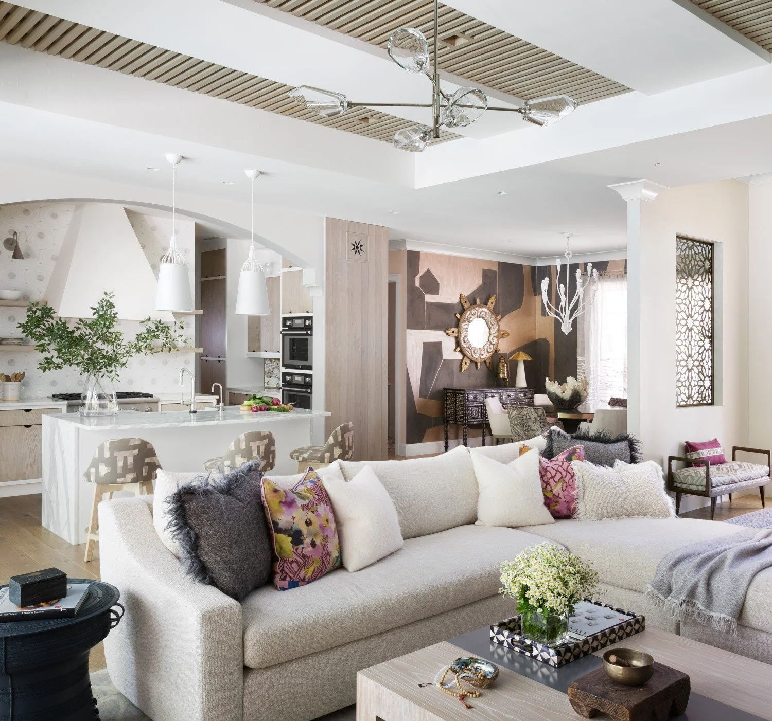 Open-concept living room and kitchen with modern decor, a white sofa with colorful pillows, a wooden coffee table with flowers, and a kitchen island with barstools.