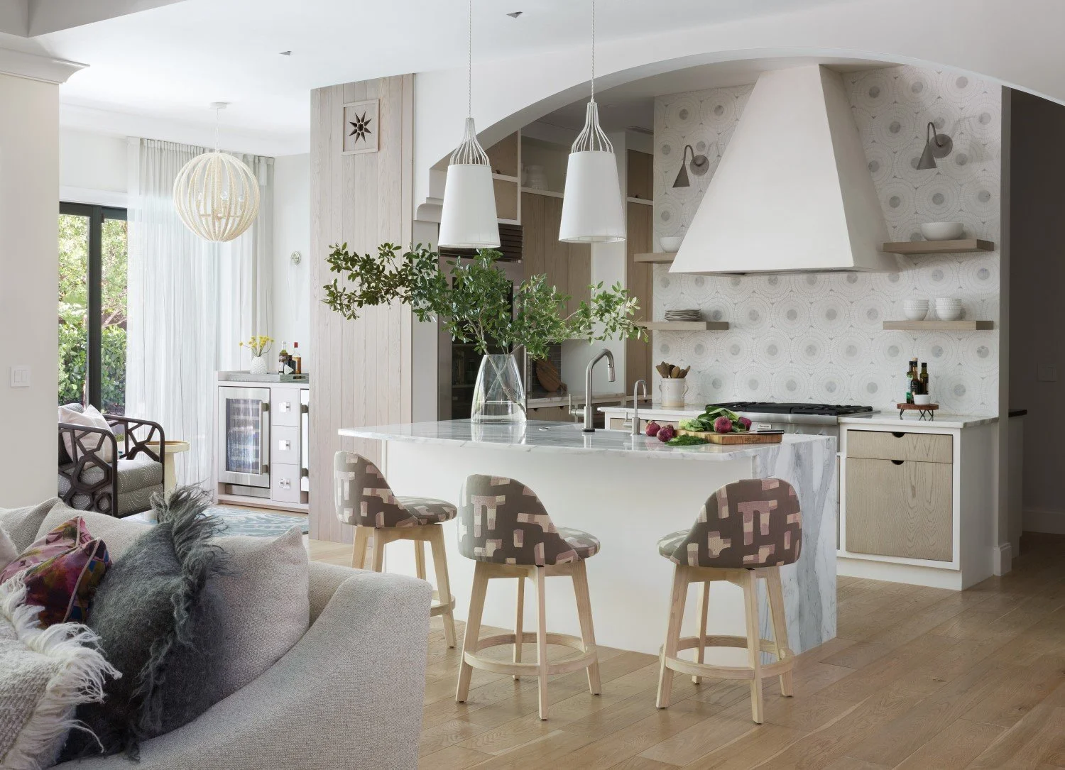 Modern kitchen in an open-plan living space with a white curved island counter, three bar stools with patterned upholstery, hanging pendant lights, a large glass vase with green foliage, and a refrigerator in the background with a small bar cart. Adj