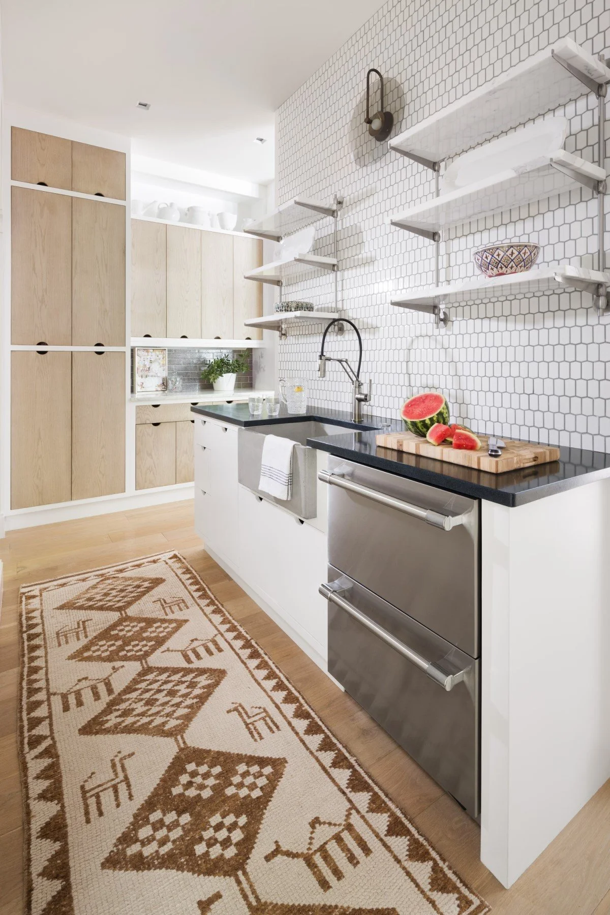 Modern kitchen with white cabinets, open shelving on a patterned tile wall, black countertop, stainless steel oven, watermelon on a cutting board, and a beige patterned rug on wooden flooring.